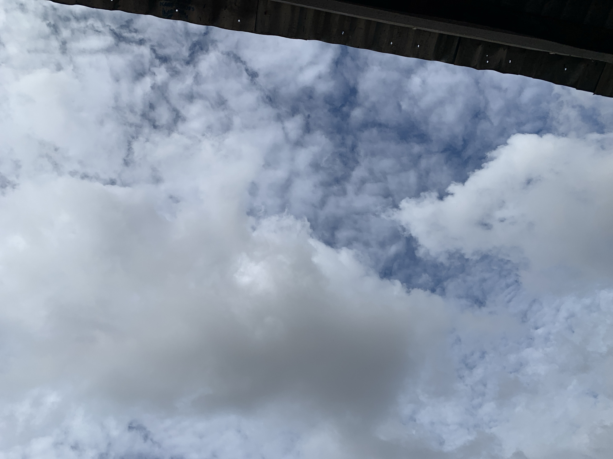 The image captures an upward perspective of a sky with mixed cloud cover, framed at the top by a dark, horizontal architectural structure. This structure, possibly an overhang or roof, displays a subtle corrugated texture and features several small, light-colored circular depressions or perforations along its visible edge. The sky is largely covered by various cumulus and stratocumulus clouds, presenting a range from bright white, indicating direct light, to softer greys where clouds are thicker or shadowed. Interspersed patches of clear blue sky are visible between the cloud formations. The scene depicts an outdoor environment under daylight conditions in New Stadium, Nigeria. No individuals, discernible actions, or additional specific objects are present within the frame.