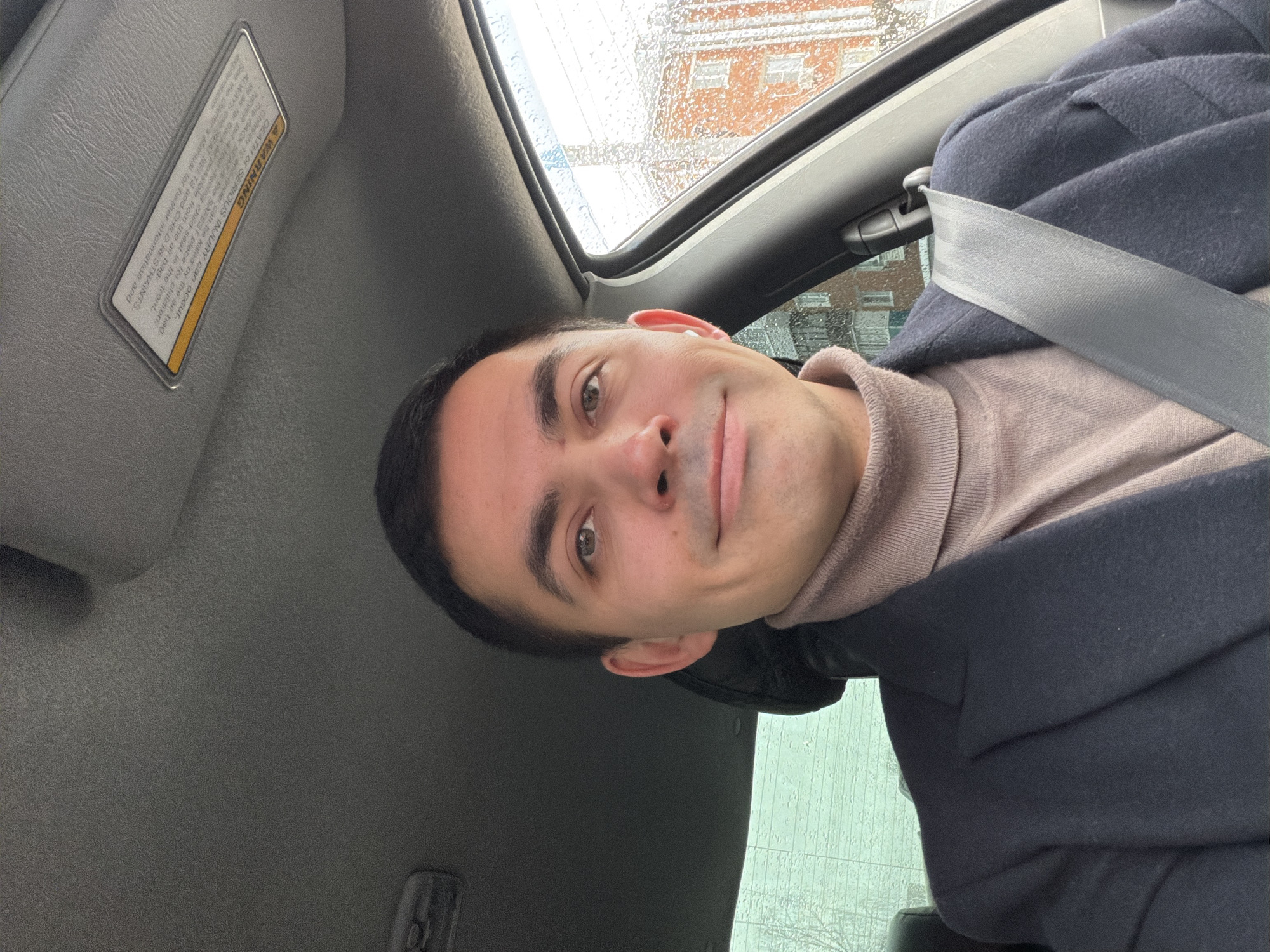 The image shows a man sitting in the driver's seat of a car. He is wearing a beige turtleneck sweater and a navy coat, with a seatbelt across his chest. The car's sun visor is down, and there is a warning label visible on it. Raindrops are visible on the car's windshield, with a blurry view of a building in the background. The location is Izhevsk, Russia.