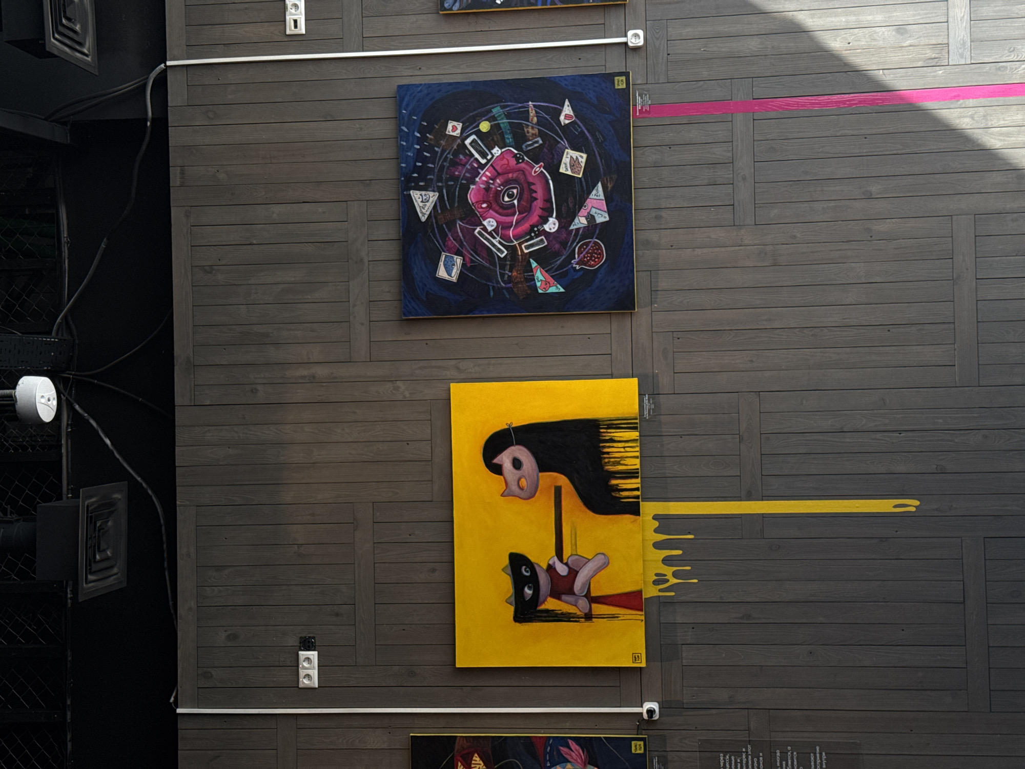 A wall in Perm, Russia, is the setting for this image. The wall, made of gray wood panels arranged in a rectangular pattern, showcases several paintings. One painting is dark blue with a pink central design and various geometric shapes around it. Another painting is yellow and depicts two cartoon-like figures, one with black hair and the other wearing a black mask. A drip effect extends from this painting onto the wall. The paintings are arranged vertically, with the yellow one below the blue one. A strip of pink paint or tape is visible on the wall above the blue painting. Metal rods are visible running horizontally across the wall, likely used for mounting the artwork. On the left side, there is a glimpse of a dark interior with metal structures. Electrical outlets are visible on the wall. The natural light casts a shadow across the wall, suggesting an outdoor or well-lit environment.