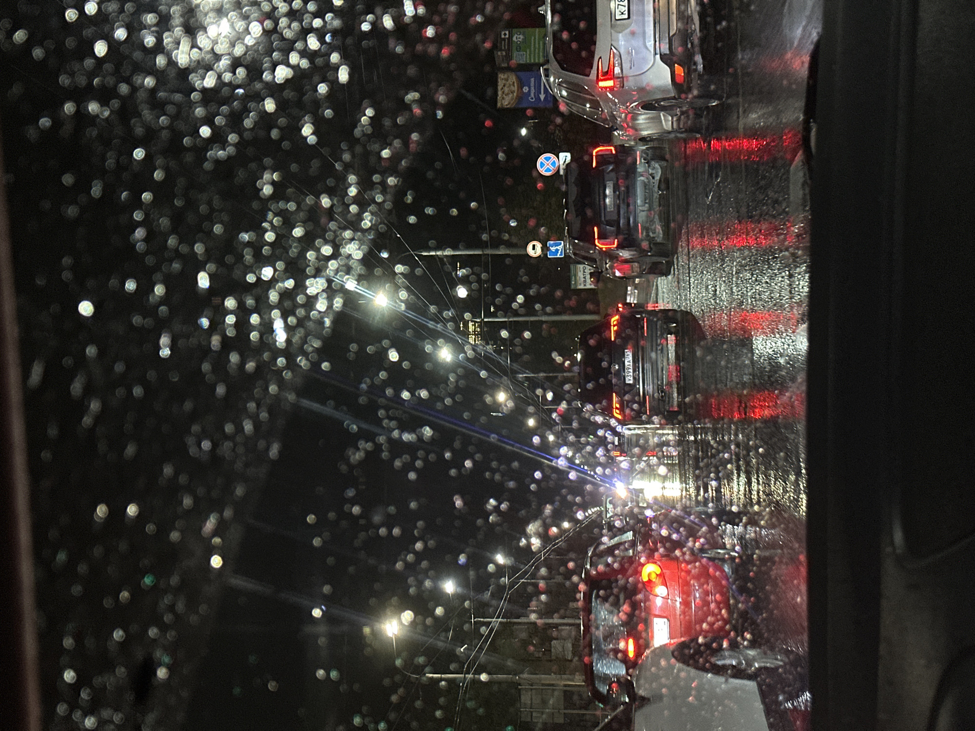 The image shows a view through a car windshield on a rainy night, capturing a street scene with several cars lined up, presumably in traffic. The windshield is covered in raindrops, which create a bokeh effect with the streetlights and car lights. The cars in front have their headlights and taillights illuminated, reflecting on the wet road surface. Streetlights are visible in the background. Based on the location (Ovoshchnoy, Russia) and the signage in the image, this is likely a street in Russia. No people are visible directly, but their presence is implied within the cars. The focus is on the immediate environment and the atmospheric conditions.