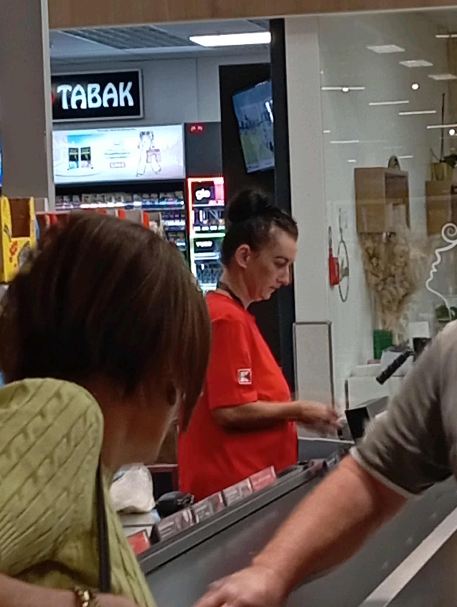 The image depicts the interior of a retail establishment, likely a grocery store or convenience store, in Nitra, Slovakia. A sign above a display reads "TABAK," indicating the sale of tobacco products. In the foreground, a person with short brown hair and wearing a light green, textured sweater is partially visible from the back. To their right, a person wearing a red shirt with a white logo on the sleeve is attending to a transaction at a checkout counter. This person appears to be a store employee. Another individual, only their arm and shoulder are visible, is interacting at the counter, suggesting a customer. The environment appears to be well-lit, with a clean and organized checkout area. The overall impression is that of a typical retail transaction taking place in a commercial setting.