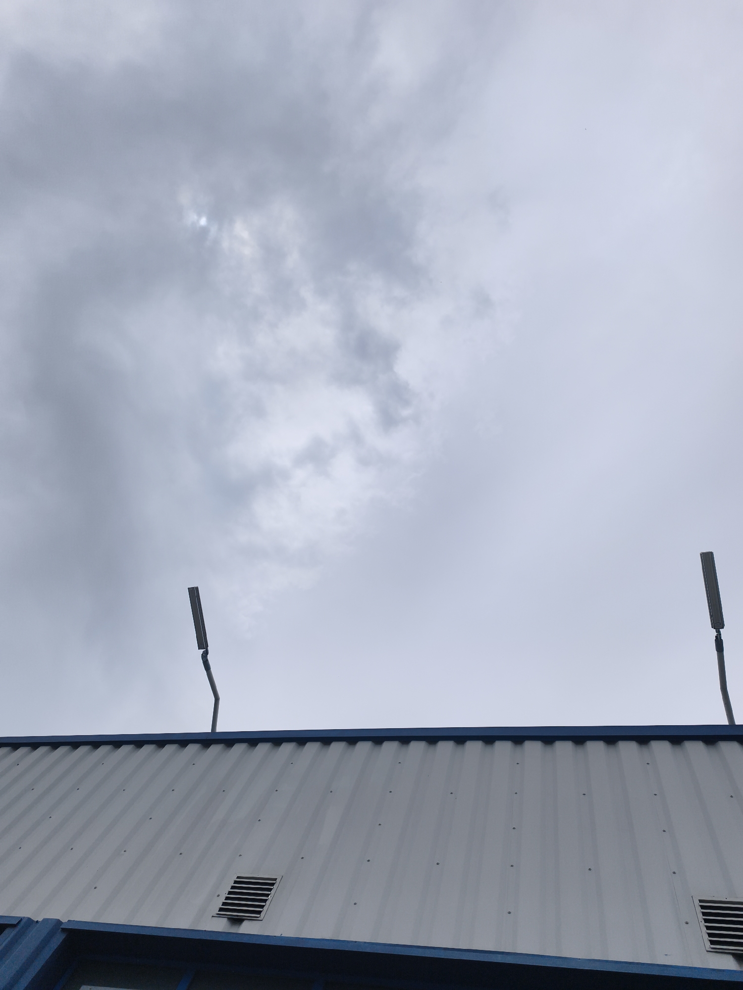 The image presents an upward view of the top section of a building set against a heavily overcast sky. The sky is dominated by thick, grey clouds, though a small, brighter patch suggests diffused sunlight attempting to penetrate the cloud cover.
The primary subject is the upper part of a building, which features corrugated metal siding or paneling in a light grey hue, secured with visible fasteners. A prominent blue trim or fascia runs along the top edge of the structure. Two dark, rectangular ventilation grates are visible on the building's side.
Extending above the building's roofline are two modern, slender streetlights or luminaires. These lights are mounted on poles and are dark in color, standing out against the pale sky and building. Their design is contemporary and minimalist.
The scene is devoid of people or any discernible activity, creating a static composition. The weather is clearly cloudy and grey, indicative of an overcast day. Based on the provided context, the image was captured in Plovdiv, Bulgaria, likely depicting an industrial or commercial area. No text is visible within the image.