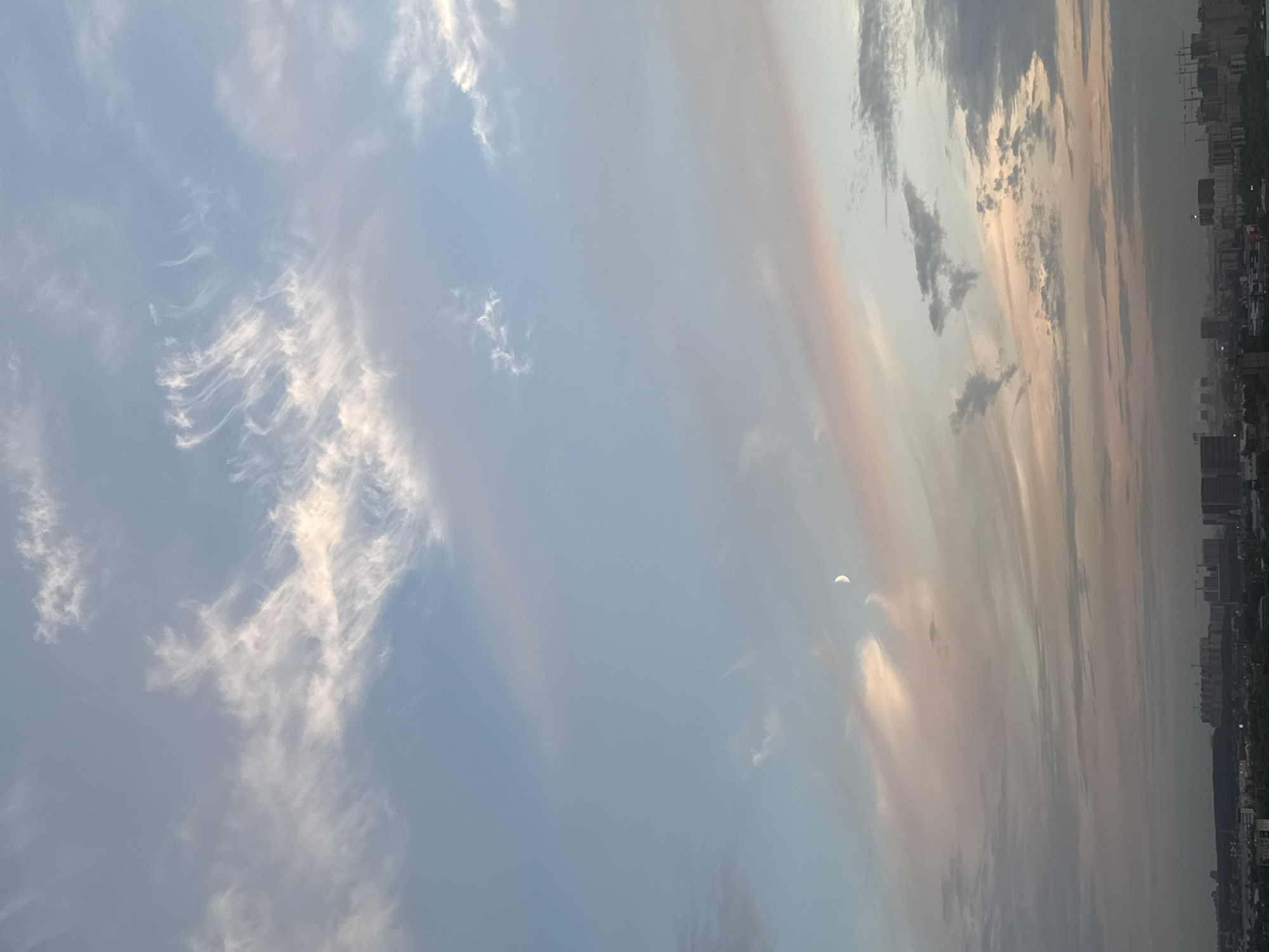 The image displays a cityscape of Lyubertsy, Russia, under a sky filled with wispy clouds at dusk. The sky transitions from a pale blue at the top to shades of pink and grey closer to the horizon. A crescent moon is visible in the upper left quadrant of the sky. Below the sky is a silhouette of the city, with various buildings of different heights and construction cranes dotting the skyline. The foreground shows a dense collection of structures and trees, suggesting a built-up urban environment. The overall impression is of a city at the end of the day, with hints of ongoing construction and development.