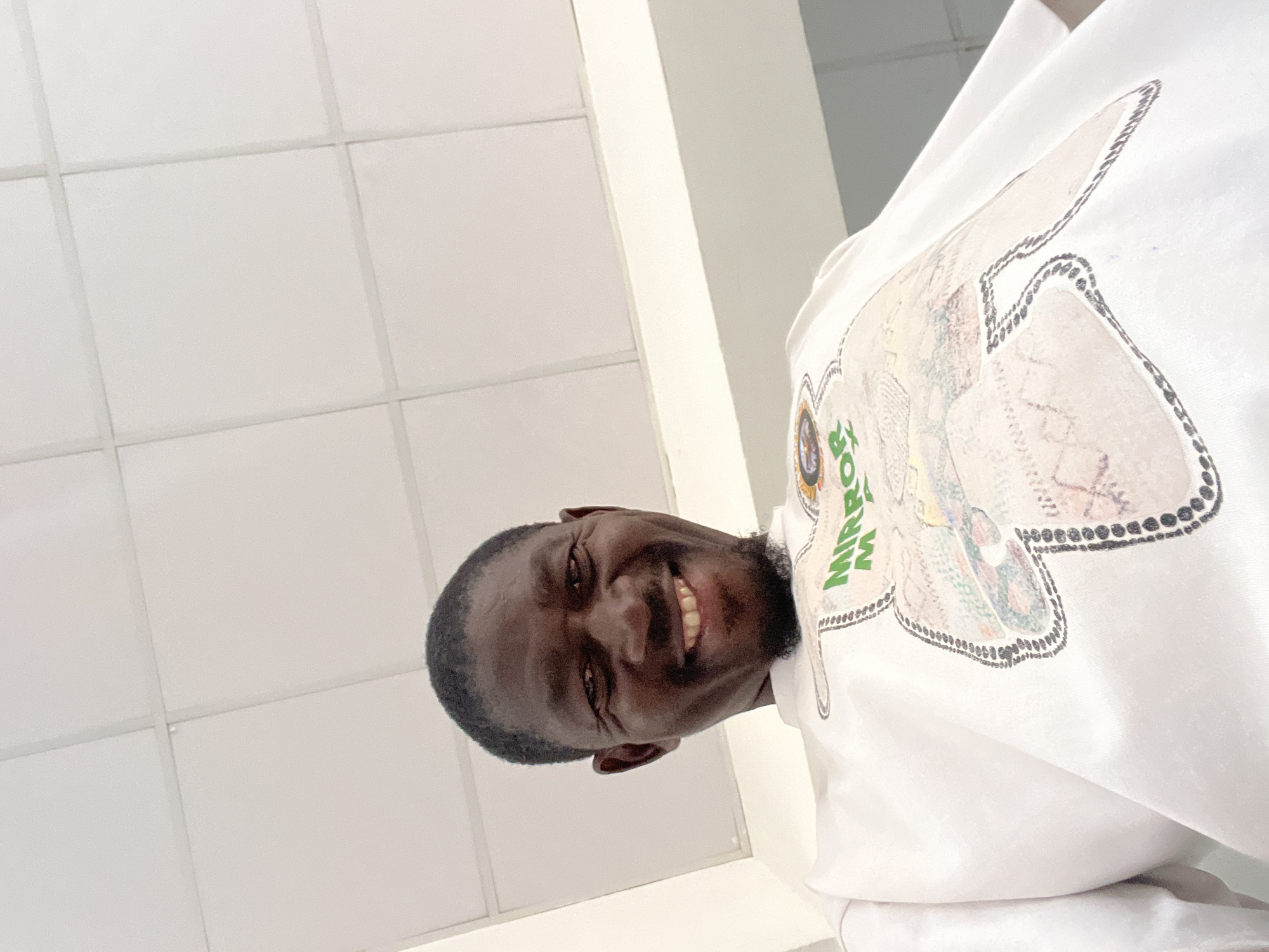 The image shows a low-angle, close-up shot of a Black man smiling broadly. He is wearing a white t-shirt with a colorful graphic design on it, which includes the words "MIRROR MA". The man has short dark hair, a goatee, and appears to be indoors, possibly in a hallway or room with a white tiled ceiling. The overall lighting suggests an interior setting. There is no information in the image to suggest a specific location in Jos, Nigeria.