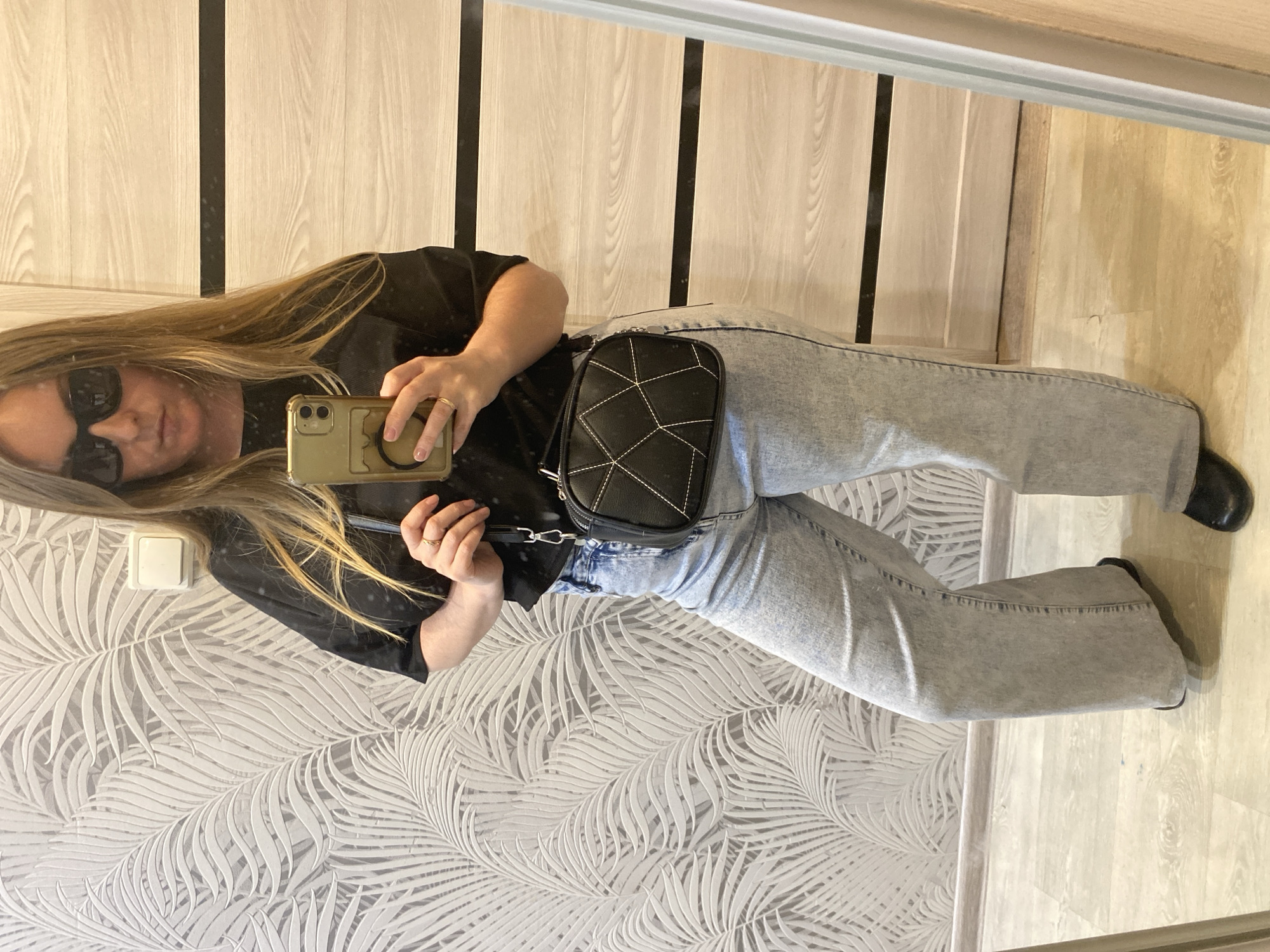 A woman is standing in an indoor setting, possibly an apartment or home interior, and taking a photo of herself in a mirror. The woman is wearing sunglasses, a black top, light blue jeans, and black shoes. She has long blonde hair. She is holding a smartphone with a light-colored case. Over her shoulder is a small black bag with a quilted design. The background includes a wall with a light-colored palm leaf pattern wallpaper and a door. The location appears to be an interior of a residence. There is no evidence to suggest the city of Noviy, Russia, from the visible content of the image.