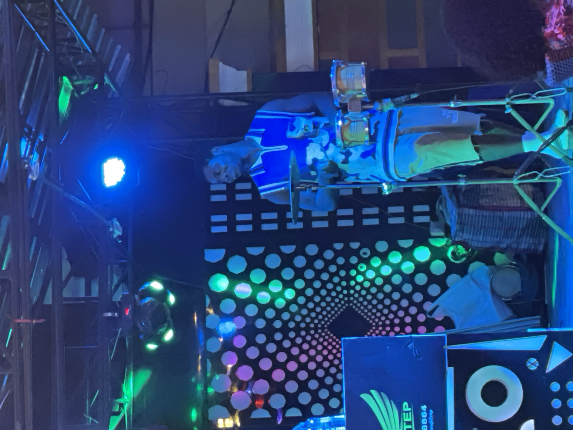 A male musician performs on an illuminated stage in Obosi, Nigeria. The dark-skinned man, with short dark hair, stands center-right, wearing a blue and white sleeveless polo shirt, light-colored shorts, and white shoes with white socks. He is positioned behind a set of bongo drums mounted on a stand, with a cymbal mounted to his left. His hands are raised above the drums, indicating he is actively playing or poised to play.
To the left, a large dark speaker box is visible, featuring a white "WING STEP" logo with a wing graphic and the number "8864". Below this, illuminated geometric shapes are present on the speaker's surface. The stage backdrop consists of a dark panel with a concentric white circle pattern, illuminated in transitioning shades of pink, purple, green, and blue. Overhead, metal trusses support various stage lights, including a prominent blue spotlight and a rotating light fixture emitting green light. To the right of the performer, exposed structural elements and wall sections of a building are visible. A portion of a second individual's head with dark, curly hair is visible in the bottom right corner of the frame. The stage floor is bathed in blue light, and miscellaneous items, including what appears to be a white bag and a woven basket, are situated behind the musical equipment.