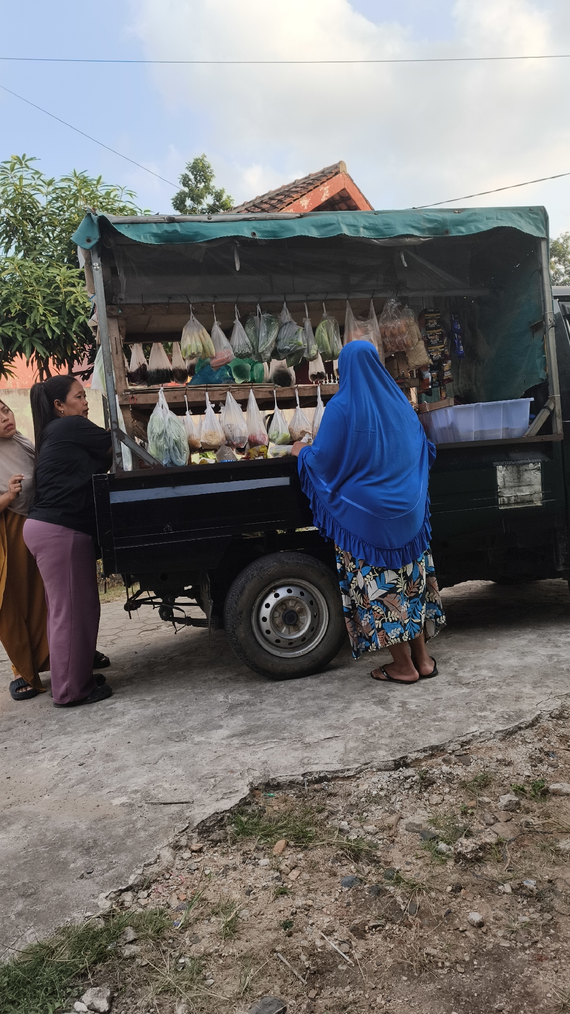 The image shows three women standing near a mobile roadside stall selling fresh produce. The stall is set up on the back of a small truck, with a green tarp for shade. Various vegetables are displayed in plastic bags hanging from a rack. One woman, wearing a blue hijab and a patterned skirt, is interacting with the vendor or selecting items from the stall. Another woman, in a black shirt and purple pants, is standing to the left, looking towards the stall. A third woman, partially visible on the far left wearing an orange skirt, is also present. The scene takes place on a concrete surface with some patches of dirt and grass. The background shows a residential building with a tiled roof and trees. The overall setting suggests a casual marketplace or street vendor scene in a neighborhood. Given the style of the stall and the people's attire, this scene is consistent with a street-side market in Bandar Lampung, Indonesia.