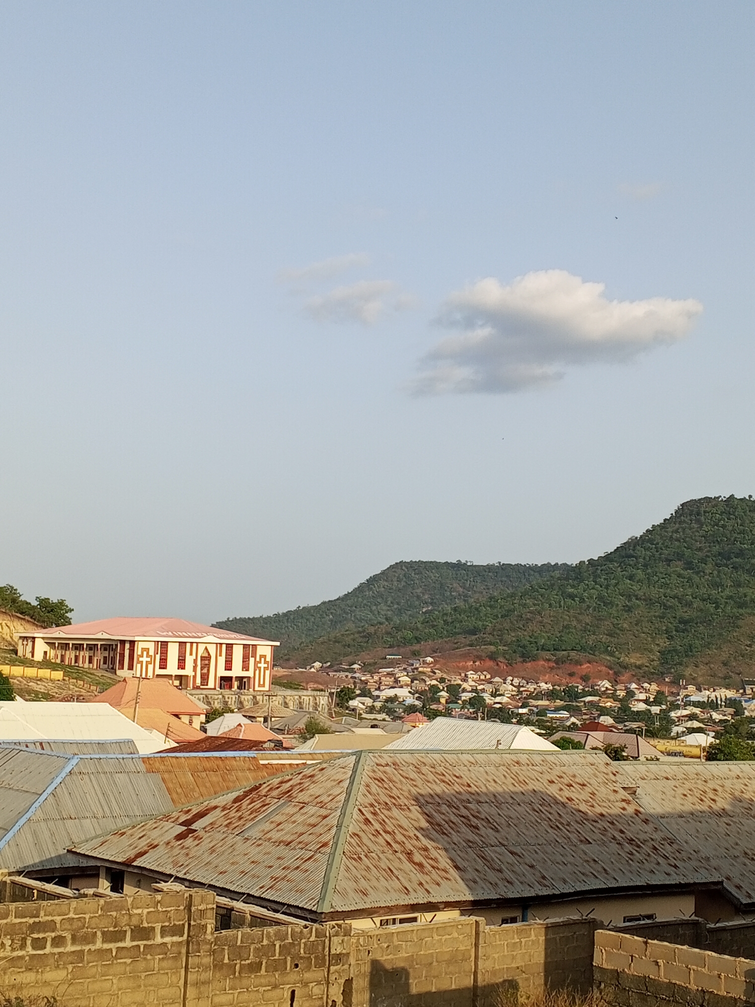 The image depicts an elevated view of a residential area in Lokoja, Nigeria. In the foreground, a partially constructed cinder block wall is visible. Beyond the wall are rooftops of corrugated metal, some showing signs of rust and weathering. A large building with a pink roof, featuring cross-shaped architectural details, stands on a higher elevation in the midground. Behind the buildings, a densely populated area of houses extends toward the base of a forested mountain range. The sky is light blue with scattered clouds. A small dark object, possibly a bird, is visible in the sky. The overall scene suggests a town nestled between hills and mountains.