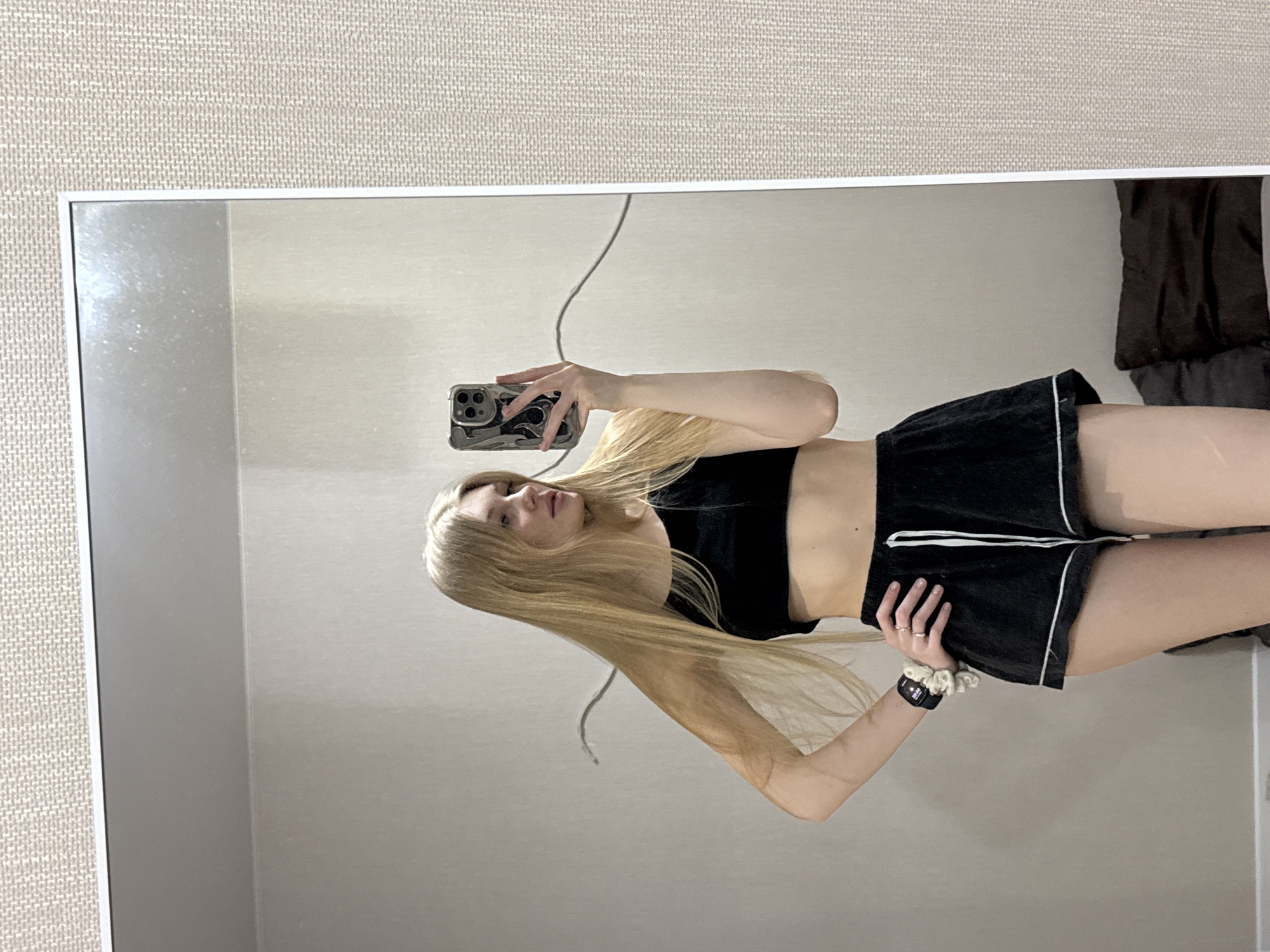 A young woman with long blonde hair is taking a selfie in a mirror. She is wearing a black crop top and black shorts with white trim. She has a black smartwatch and a white scrunchie on her left wrist. The location appears to be an indoor setting with a light-colored wall. The image does not provide information about the city of Perm, Russia.
