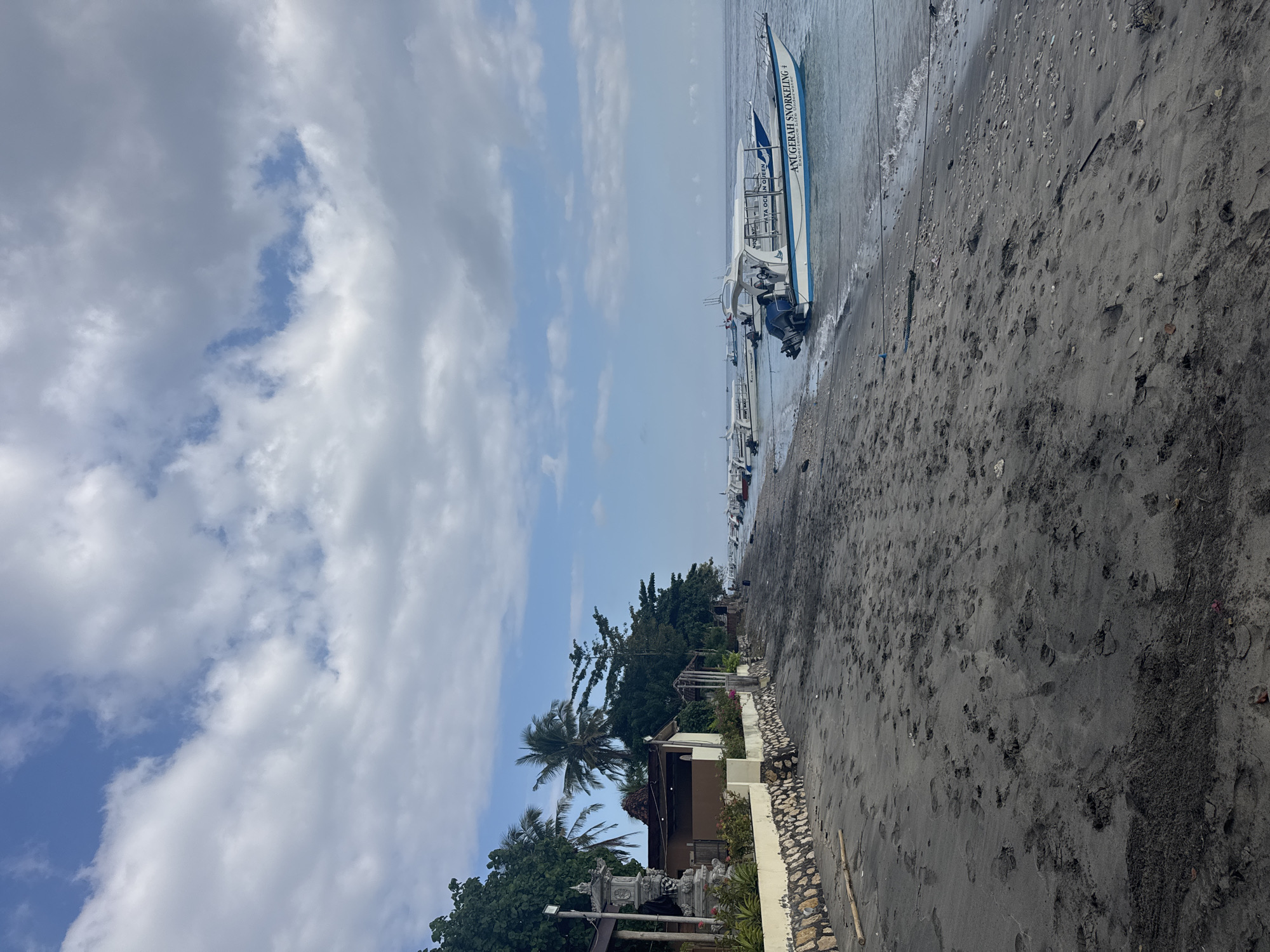 The image depicts a tropical beach scene in Nusa Penida, Indonesia, with a focus on the coastline and the water. The beach itself is composed of dark sand, marked with numerous footprints, suggesting recent human activity. A low, stone-and-plaster wall runs along the edge of the beach, separating it from a line of trees and buildings. These structures appear to be small bungalows or shops, with palm trees and other lush vegetation providing shade.
Several boats are anchored in the shallow, calm water just offshore. The most prominent boat in the foreground is a white and blue vessel with an open passenger cabin, featuring the text "ANUGERAH SNORKELING" and "Experience The Underwater" on its side, indicating it is likely used for tours. Other similar boats are visible further down the coast.
The sky above is partly cloudy, with patches of blue sky interspersed with fluffy white and grey clouds. The lighting suggests it is daytime, possibly late morning or early afternoon. The overall atmosphere is tranquil, characteristic of a popular tourist destination.