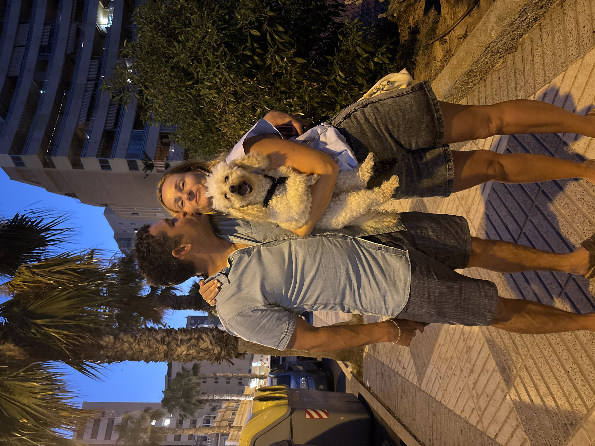 A man in a blue denim shirt and grey shorts kisses a woman on the cheek. The woman, wearing a light blue shirt and denim shorts, holds a small, fluffy white dog. The dog has its mouth open, as if panting or barking. They are standing on a sidewalk made of light-colored rectangular pavers, with a curb to their right. Behind them are palm trees, a building with balconies, and a street with a parked car. To the left, there are two large, grey trash bins with yellow lids. The scene is lit by artificial lights, casting long shadows on the sidewalk. The overall impression is a casual moment captured outdoors at dusk or early evening in Calp, Spain.