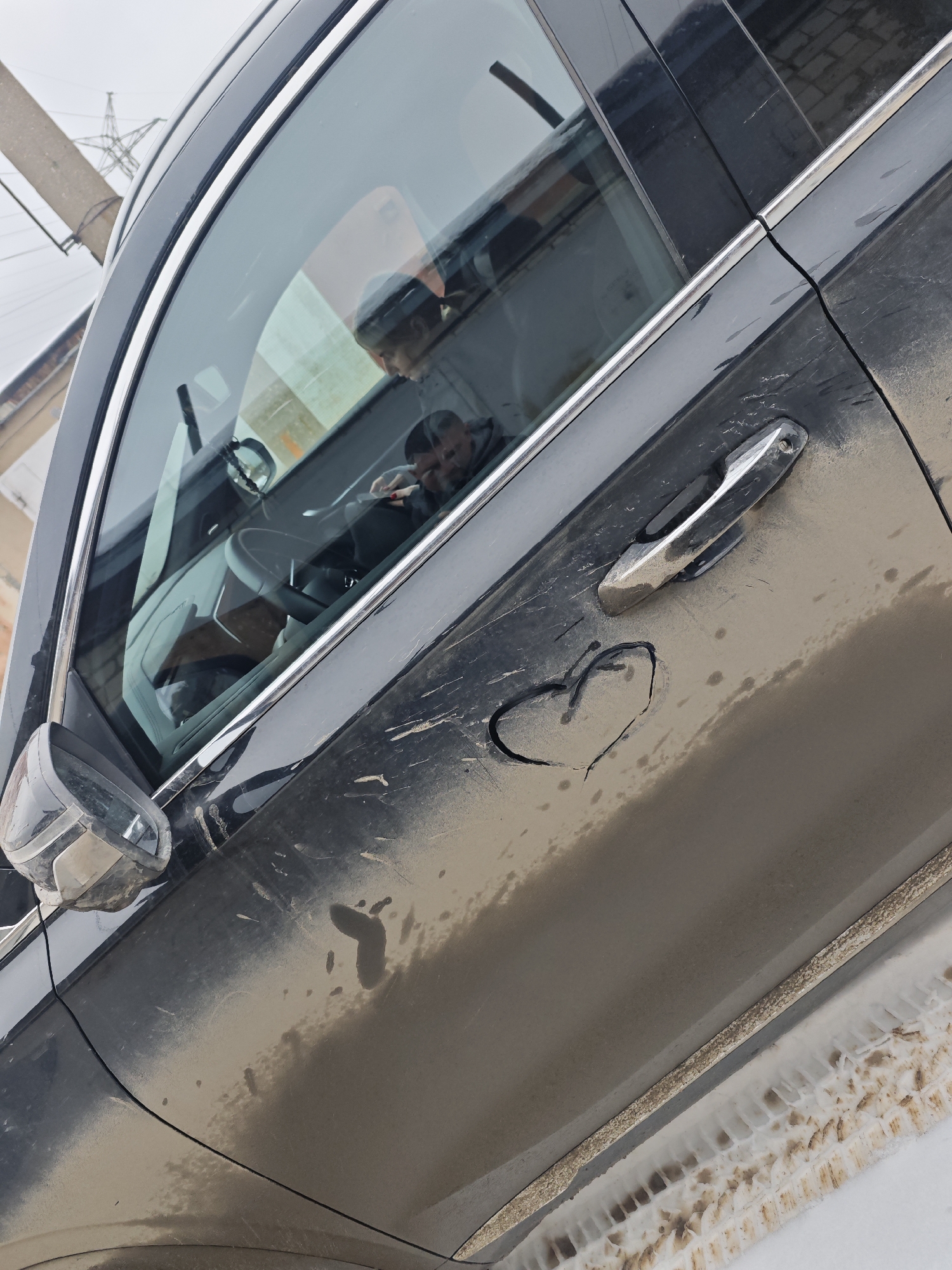 The image shows a black SUV parked on a street in the city of Chaikovsky, Russia. The car is covered in a layer of dirt and grime, with a heart shape drawn in the dust on the driver's side door. A person is visible in the driver's seat looking at something in their hand. Snow and slush are visible on the ground near the car's tires. Power lines and a brick building can be seen in the background.