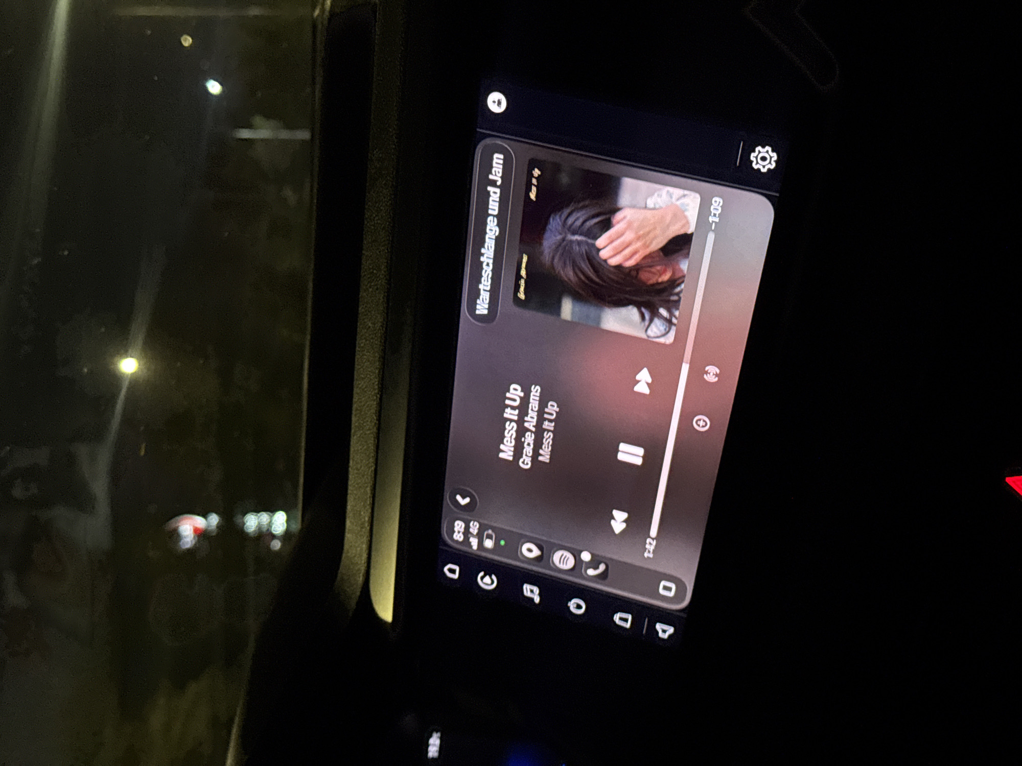 Here is a forensic description of the image:
The image shows a car's infotainment screen at night. The screen displays the Spotify music player. The song playing is "Mess It Up" by Gracie Abrams. The music player also shows the "Warteschlange und Jam" queue. The time left on the song is -1:09. The screen also shows the phone's data is connected to 4G and the time is 8:19. Outside the car is dark and blurred, indicating the vehicle is likely in motion at night. The location and specific environment are not discernible.