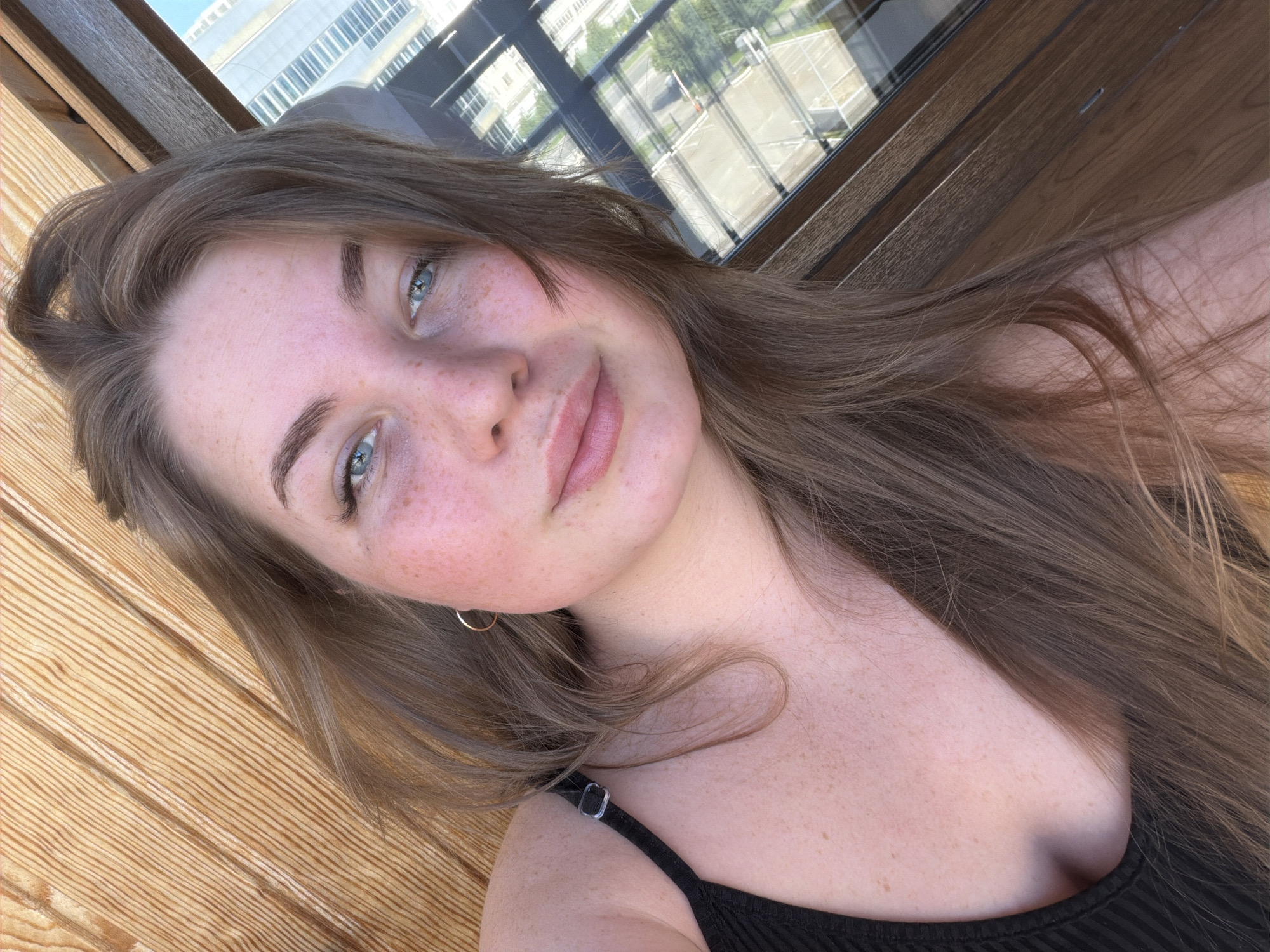 A light-skinned female, appearing to be a young adult, is depicted in an upward-angled selfie taken in the city of Kazan, Russia. She has long, light brown hair, which is somewhat tousled, and blue eyes. Her face shows numerous freckles across the nose and cheeks, and her lips are full with a light pink tint. She is wearing a black ribbed tank top, with a silver-colored strap adjuster visible on her left shoulder. A small, thin gold hoop earring is present in her right ear.
The background to the left features light brown wooden paneling with distinct parallel grain patterns. To the right, a large window with a dark brown wooden frame offers a view of a cityscape under daylight. Through the window, multi-story buildings, a paved road with what appears to be a single vehicle, and green trees and foliage are visible. The scene suggests an indoor or sheltered outdoor setting like a balcony, overlooking an urban environment.