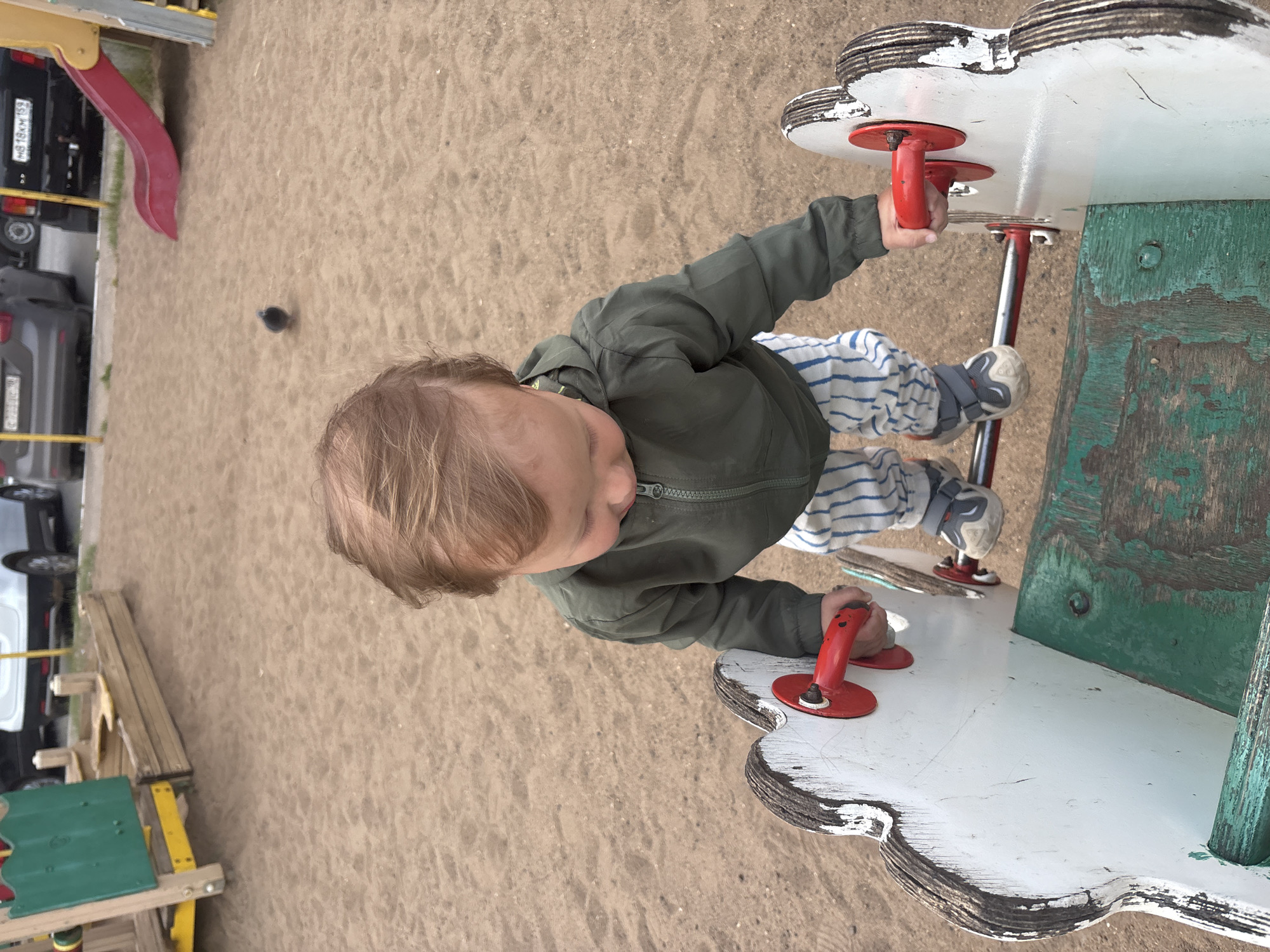 A young child, wearing a green jacket and striped pants, stands on a wooden rocking toy at a playground. The child is holding onto red handles on the toy. The ground is covered in sand. In the background, there is a slide and a sandbox, as well as parked cars. The scene appears to be in the city of Chaikovsky, Russia.