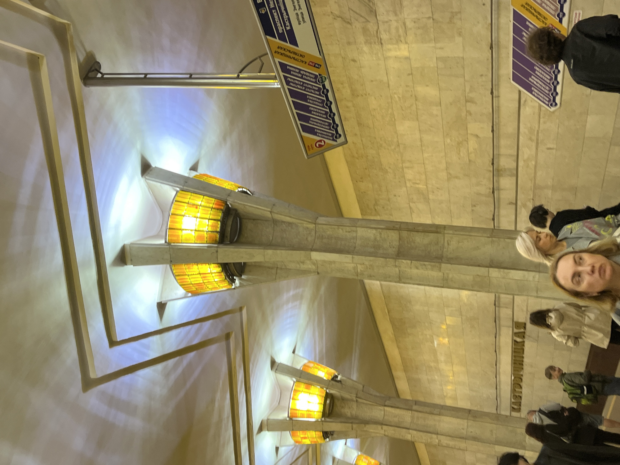 The image depicts the underground platform of the Kastrychnitskaya (Oktyabrskaya) metro station in Minsk, Belarus. The scene features multiple concrete pillars, each topped with a distinct circular light fixture enclosed by amber-colored translucent panels, emitting upward-directed illumination onto a white ceiling. The ceiling exhibits rectangular recessed panels and reflects both the warm yellow light from the fixtures and cooler blue-white light, creating intricate shadow patterns. The platform walls are clad in light-toned, horizontally laid stone or tile.
A metro information sign, displaying "Line 2" (purple line) and a series of station names in Cyrillic, including "КАСТРЫЧНІЦКАЯ" and "ОКТЯБРЬСКАЯ," is suspended from the ceiling on the right side of the frame.
Approximately seven individuals are present on the platform. In the immediate foreground, a woman with blonde hair and a neutral expression looks slightly to her left. To her right, another woman with blonde hair faces towards the right side of the frame. Further back, near the wall, a person with a green backpack is visible, looking left and exhibiting a slight smile. A person in a light beige jacket is partially visible, facing away from the viewer. On the far right, a person with dark hair, wearing a dark jacket, is observed from behind, standing near the metro information sign. Other individuals are partially visible or in the background, appearing to be waiting or transiting through the station.