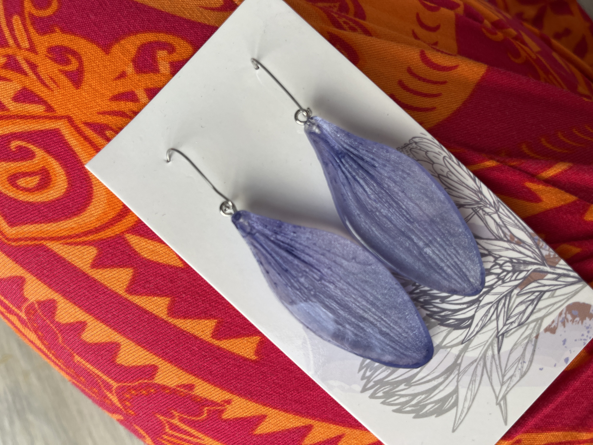The image shows a pair of purple petal-shaped earrings displayed on a white card. The card has a floral illustration printed on it. The earrings are made of a translucent material and have delicate vein-like details. They are suspended from silver hooks. The background consists of a vibrant fabric with a pattern of alternating pink and orange stripes and abstract designs. The scene appears to be a close-up shot, possibly taken indoors. The location in Сеніцкі сельскі Савет, Belarus is not determinable from the visual information provided.