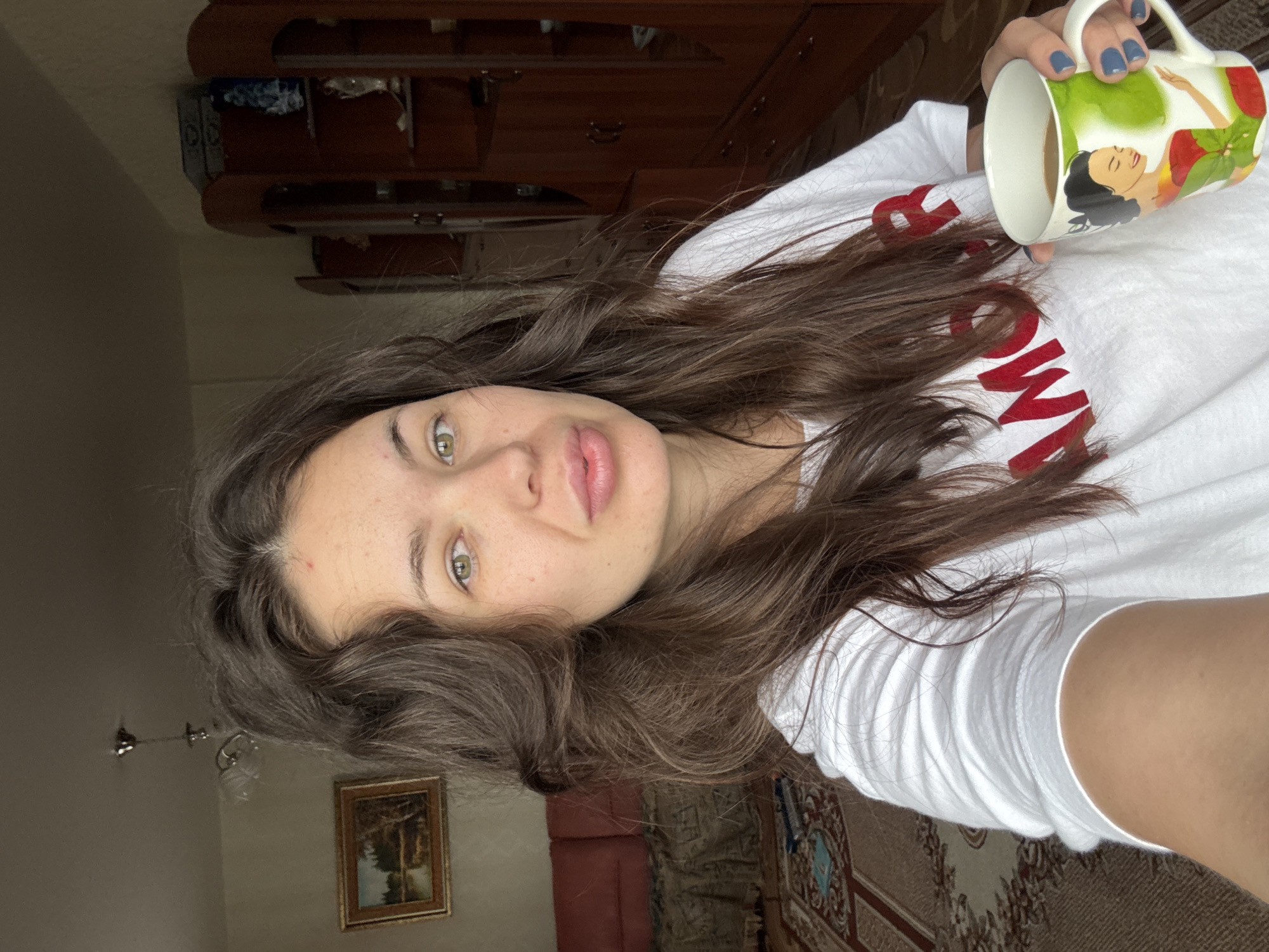 A young woman with long brown hair and freckles is holding a mug with a green and white design in her right hand. She is wearing a white t-shirt with red lettering. The background includes a brown wooden dresser, a painting on the wall, a rug, and a glimpse of a red couch. The location cannot be confirmed as Bely Yar, Russia based on the provided image.