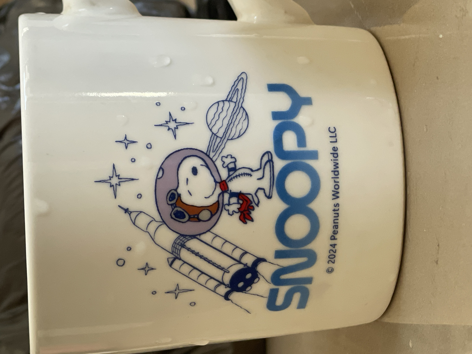 The image is a close-up, eye-level shot of a white mug with a cartoon illustration of Snoopy in a space suit. Snoopy is standing next to a rocket and a planet with rings. The word "SNOOPY" is printed in large blue letters across the bottom of the illustration, with a copyright notice for "© 2024 Peanuts Worldwide LLC" below it. The mug has water droplets on its surface. The background is dark and out of focus, suggesting the mug is placed on a reflective surface. The scene is indoors, and the lighting is natural. There is no indication of the location within Tashkent, Uzbekistan.