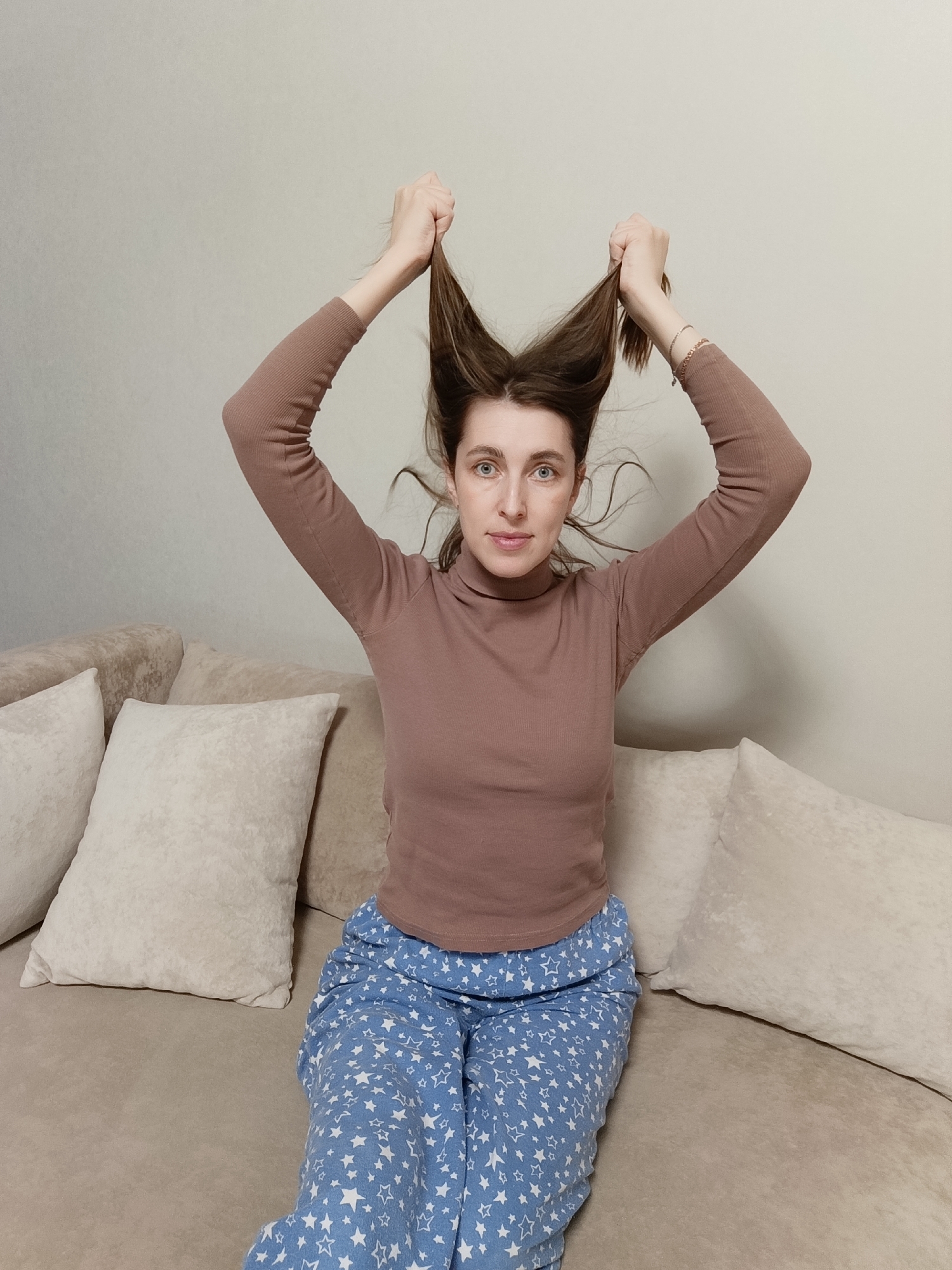 A woman is sitting on a beige sofa with several cushions. She is wearing a brown turtleneck sweater and blue pajama pants with white stars. She is holding two sections of her brown hair up above her head, creating a horn-like appearance. The background is a plain beige wall. The location is Chaikovsky, Russia.