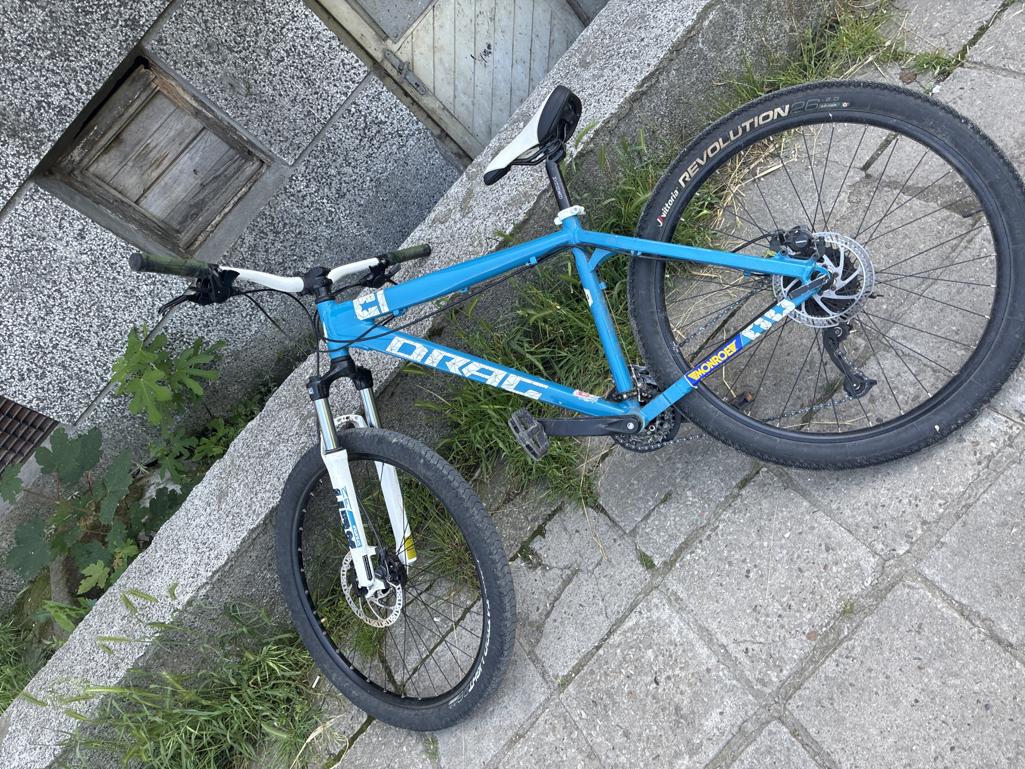 A bright blue mountain bike, branded "DRAG" on its frame, lies on its side in an outdoor setting in Varna, Bulgaria. The bike features a white seat with "XLC" visible on the seat post, and white handlebars with a mix of green and white grips. Both the front and rear wheels are fitted with disc brakes. A white "FOX" branded suspension fork is visible on the front. The rear tire is a "Vittoria REVOLUTION 26 x 2.0," while the front tire has "LITE" text visible. A "MONROE" sticker with a blue floral pattern is affixed to the chainstay.
The bike rests on a ground surface made of square concrete pavers, with green grass growing between the cracks and along the edges. Some pavers appear aged and worn. In the background, to the upper left, an old, weathered wooden hatch is embedded in a raised concrete structure. Further back, another wooden panel, possibly part of a building, is visible. Green vegetation, including some broad-leafed plants, thrives around the concrete and wooden elements. The scene is brightly illuminated, indicating a clear daytime setting. No individuals are present, and the bike is stationary, suggesting no activity is currently occurring.