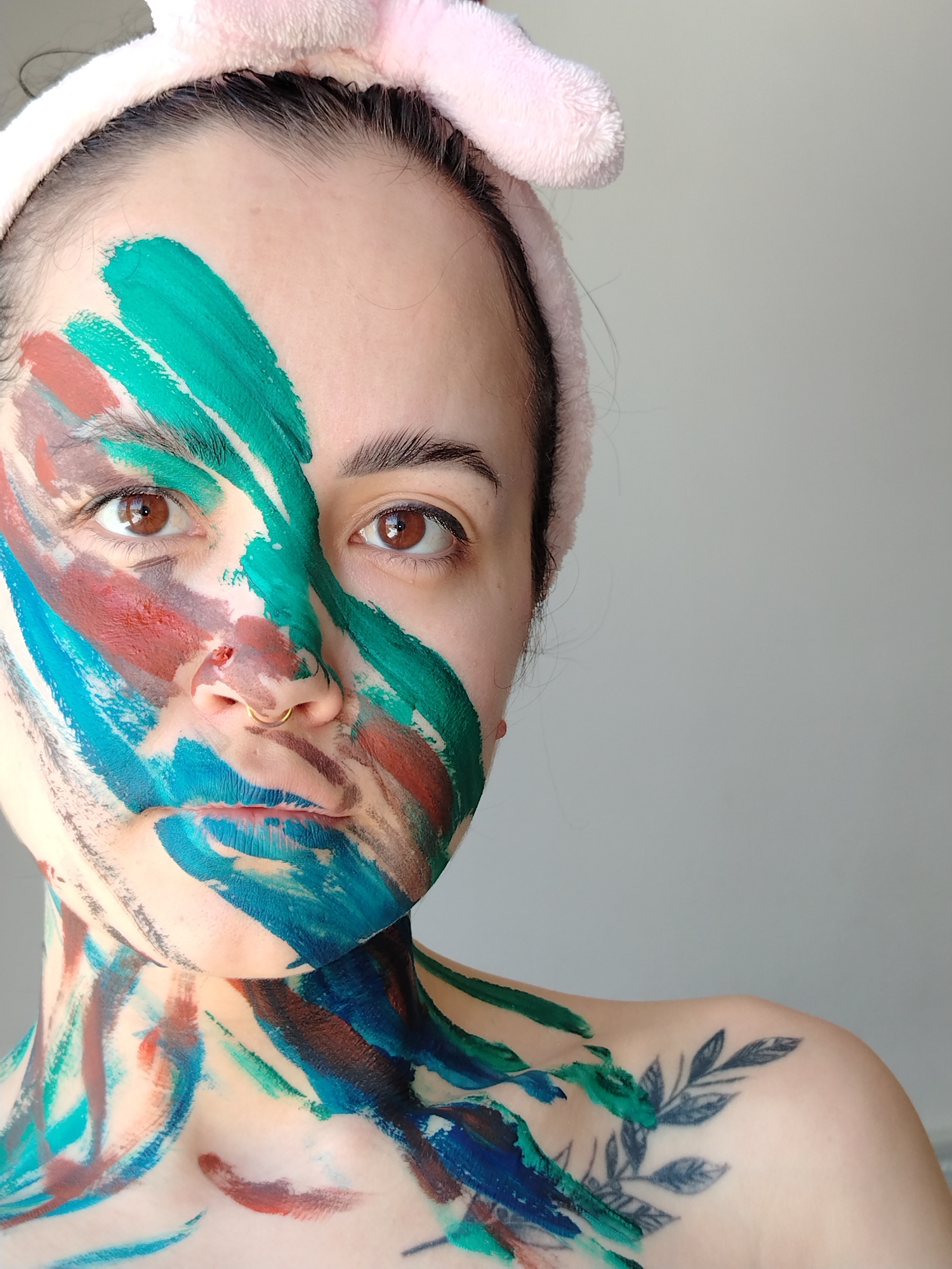 A close-up shot of a woman with paint smeared across her face and upper body. The paint consists of blue, green, and red streaks. She has brown eyes and wears a pink headband. A tattoo of leaves is visible on her right shoulder. The background is a plain light gray. There is no evidence to connect the scene to the city of Noviy, Russia.