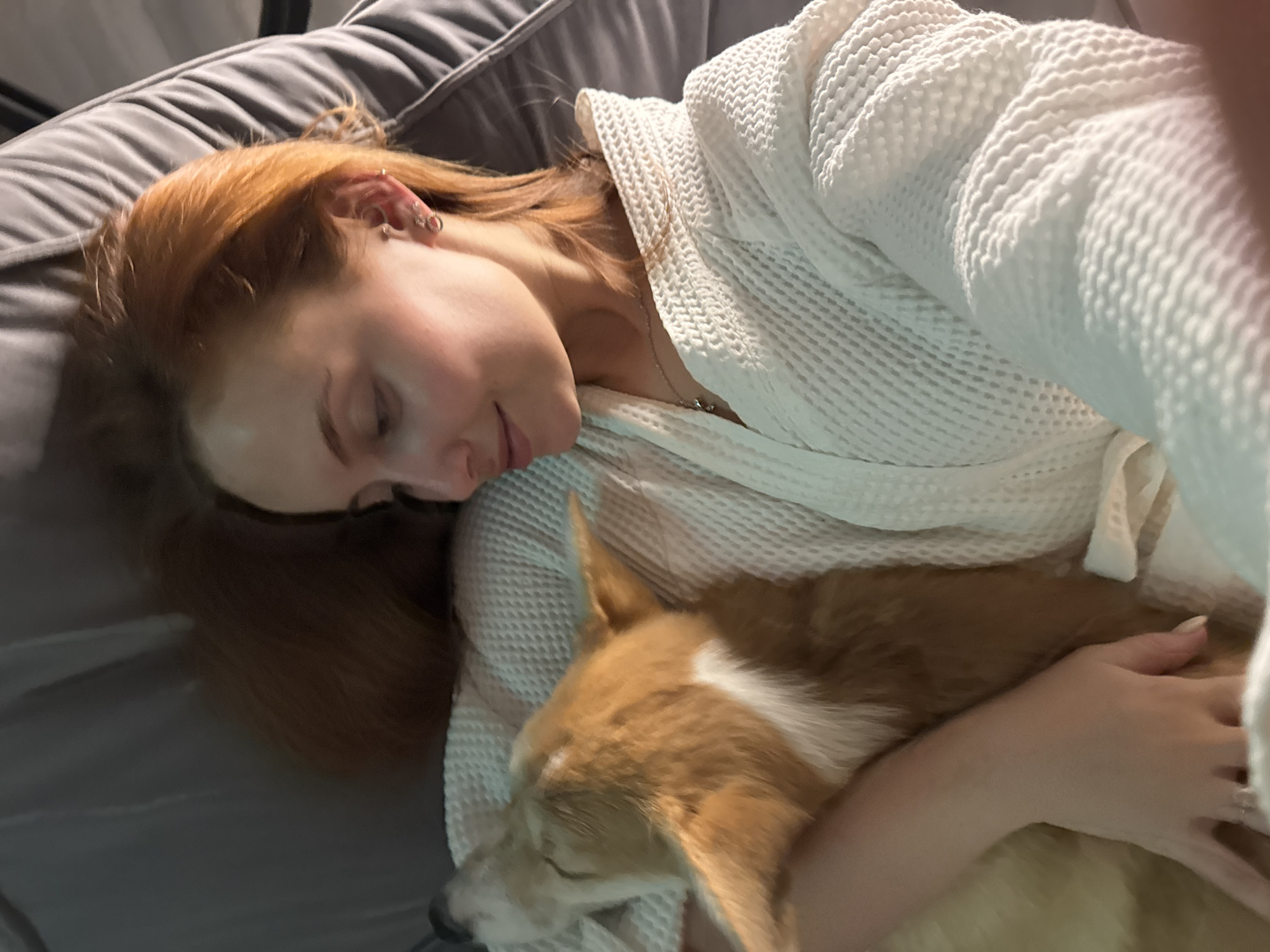A woman with long red hair and multiple ear piercings is lying on a gray couch or bed, wearing a white waffle-textured bathrobe. She is embracing a tan and white corgi dog, with her arm wrapped around it. The dog is resting peacefully with its eyes closed. The location is not discernible as being Chaikovsky, Russia, based on the image alone.