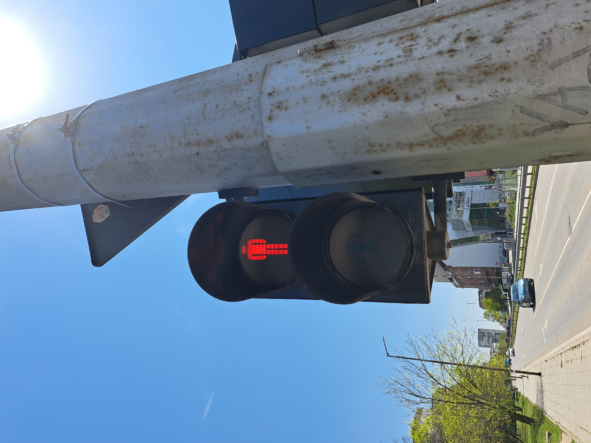 This image presents an urban street scene in Sofia, Bulgaria, captured on a bright, clear day, likely during spring or early summer due to the fresh green foliage. The perspective is angled upwards, dominated by a large, light-colored overhead gantry or pole that exhibits signs of rust and wear. Attached to this structure is a pedestrian traffic light, prominently displaying a red, stationary human figure, signaling "do not walk" for pedestrians.
Below the overhead structure, a multi-lane asphalt road extends into the distance, with several vehicles visible, including a dark car in the mid-ground. A paved sidewalk runs along the right side of the road, bordered by a strip of grass and trees with vibrant green leaves. In the background, various buildings of different sizes and architectural styles line the street. Partial text "BAR" is visible on the facade of one of these buildings. The sky is a clear, bright blue, with the sun causing a strong glare in the upper left portion of the frame, indicating daytime. The scene depicts a functional urban environment with active vehicular traffic and pedestrian control systems.