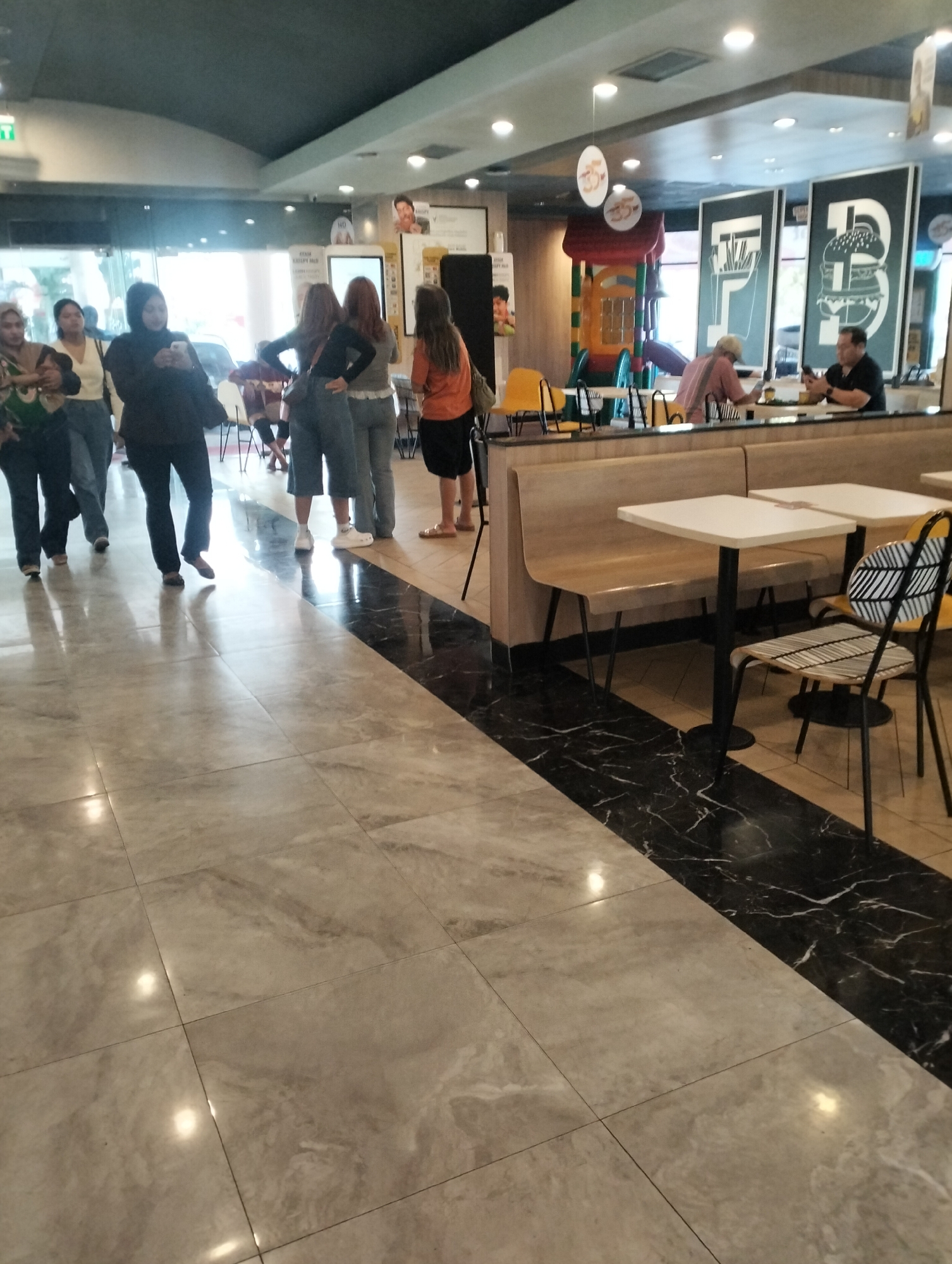 The image depicts the interior of a fast-food restaurant, likely a McDonald's, as indicated by the branding and play area visible in the background. The setting is indoors, with a tiled floor, booths, and tables. Several patrons are present, some standing in line, while others are seated at tables. The lighting appears to be artificial, contributing to the indoor atmosphere. The overall scene suggests a casual dining environment. The city of Semarang is mentioned in relation to this scene, implying this is a photograph taken within a McDonald's establishment located in Semarang, Indonesia.