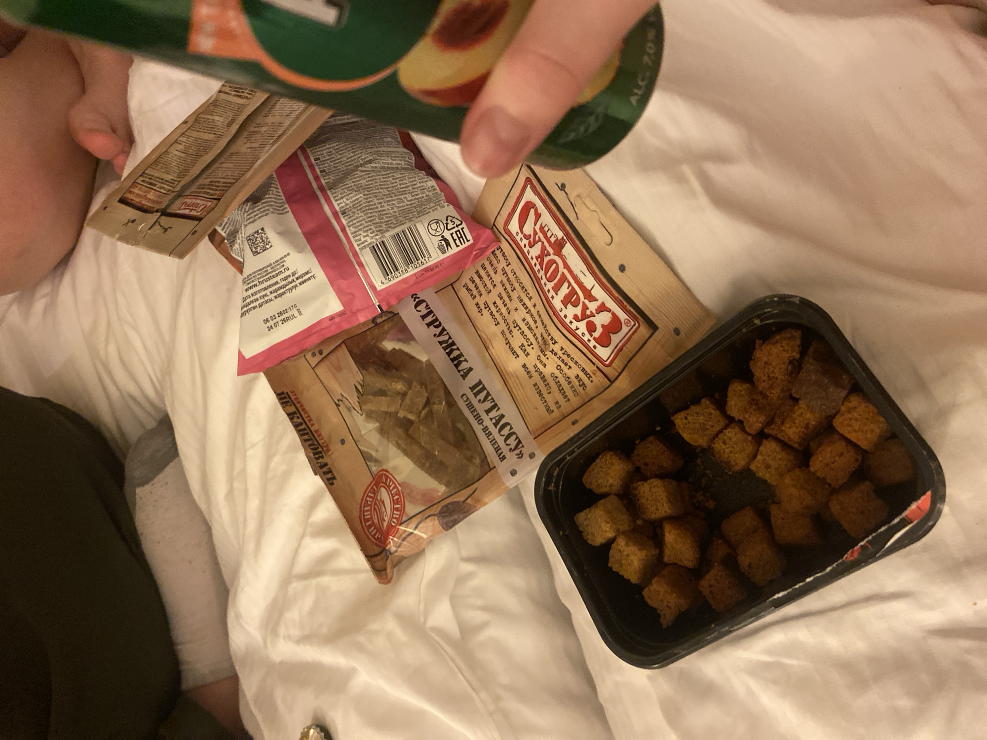 A top-down view shows an assortment of snacks scattered on a white bedsheet. The snacks consist of dried fish (branded as "Struzhka Putassu"), croutons ("Sukhogruz" brand), and accompanying condiment packets. A hand is visible holding a green and orange can of a beverage. Also visible are bare feet, a leg covered in gray fabric, and a portion of black fabric. No information related to the city of Saint Petersburg, Russia, is discernible from the image.
