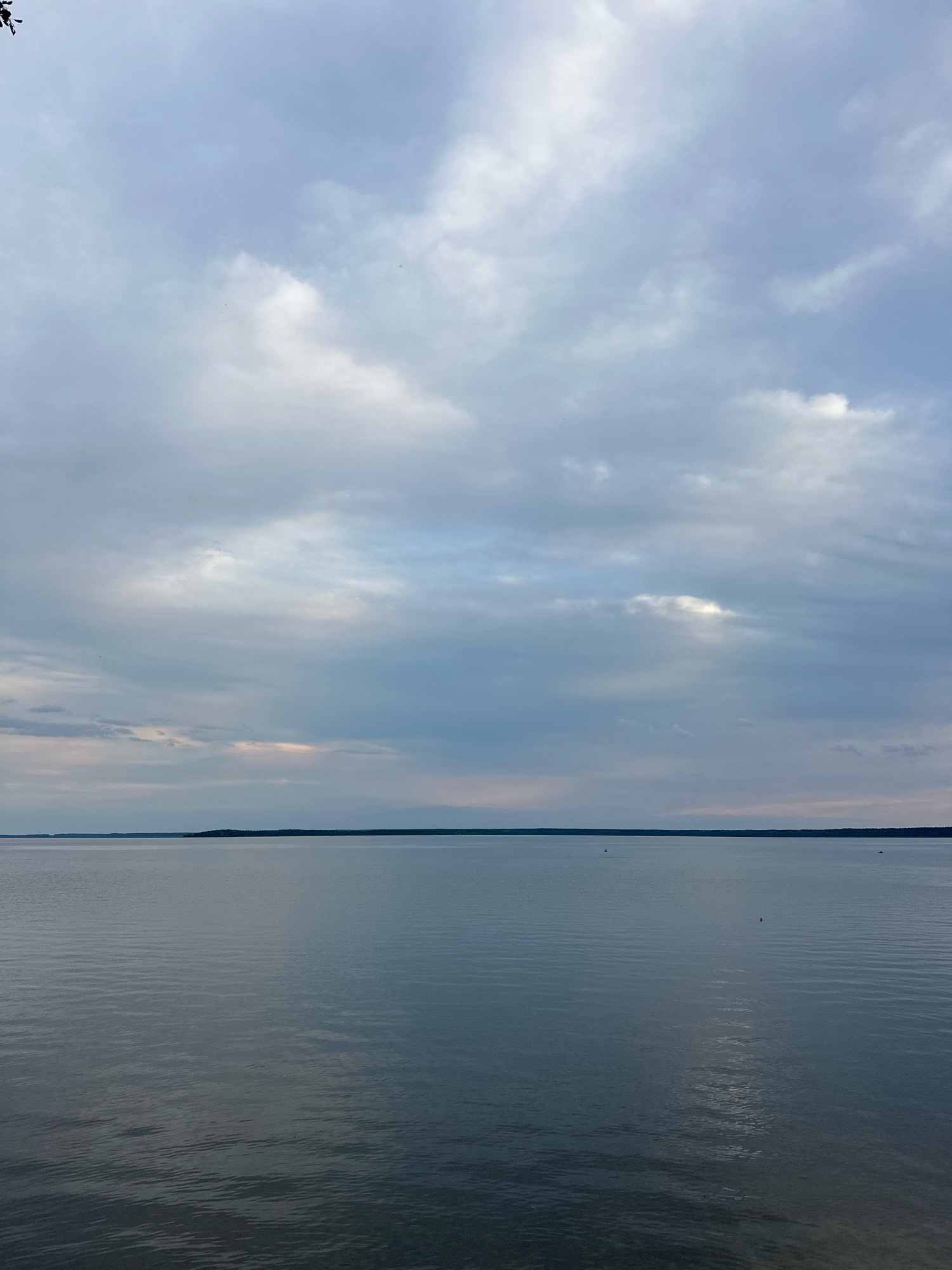 The image shows a wide expanse of calm water under a cloudy sky. The horizon is a thin, dark line separating the water from a distant, tree-lined shore. The water is a deep, muted blue-gray, with subtle ripples creating a textured surface. The sky is filled with a mix of light and dark clouds, with some patches of lighter sky suggesting the presence of sunlight filtering through. There are a few small boats visible on the water, appearing as dark specks. The overall impression is one of tranquility and open space. The location is the Narač Lake in the Narač rural council of Belarus.