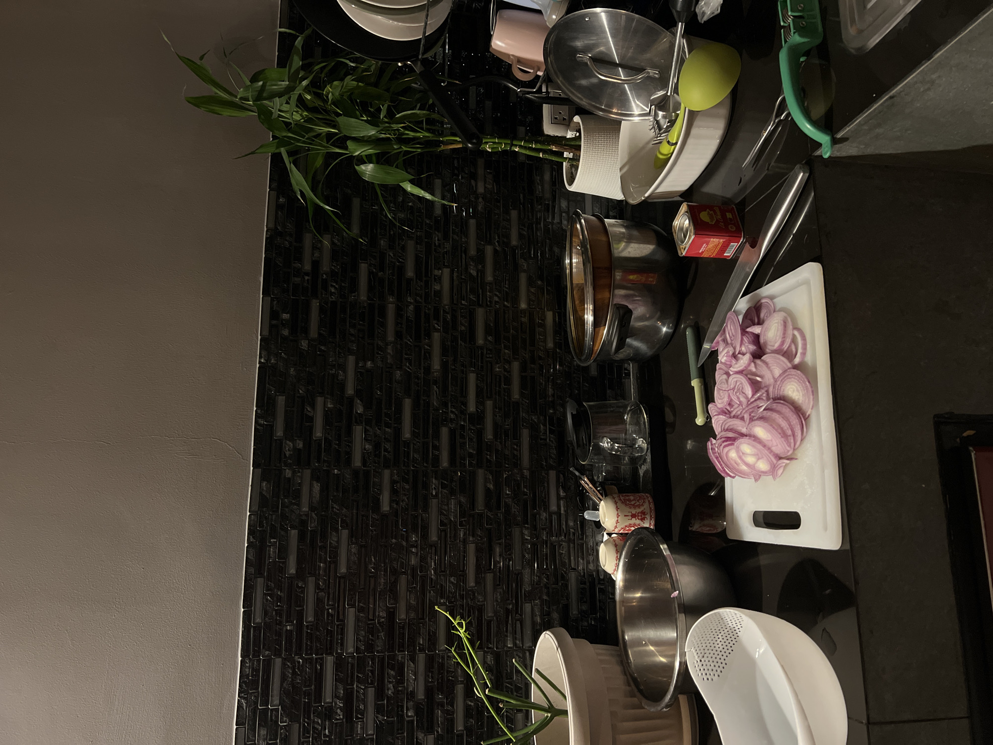 The image shows a kitchen counter with various cooking items and ingredients. A chopping board with sliced red onions sits next to a large knife. There are several pots and pans, some stacked, as well as a colander and other cooking utensils. A plant in a white pot adds a touch of greenery. The backsplash behind the counter features a dark, textured tile pattern. The overall setting appears to be a domestic kitchen environment.