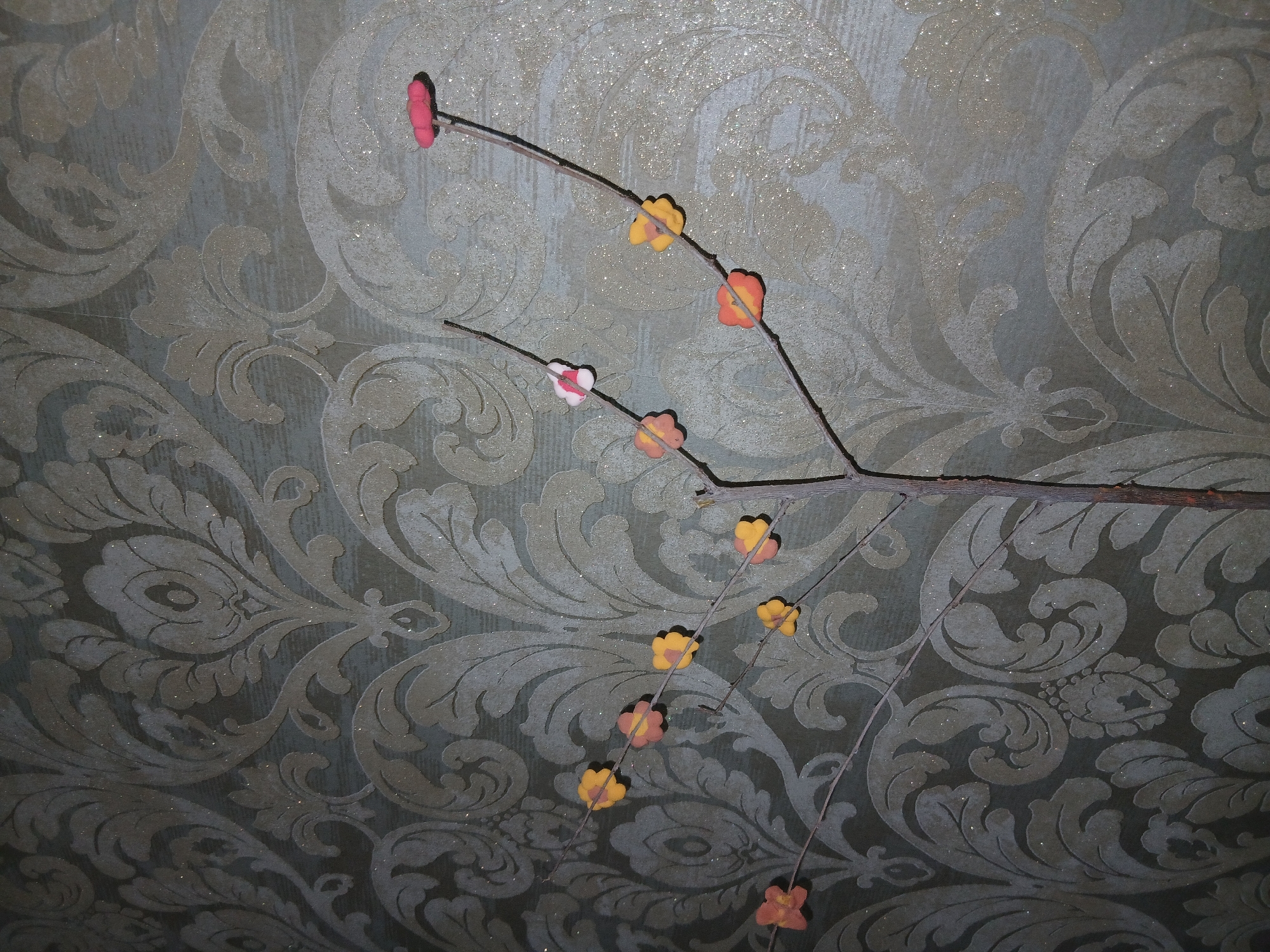 A close-up, vertical shot displays decorative branches with colorful, flower-shaped embellishments against a patterned wallpaper. The wallpaper features a damask design in muted tones of grey and teal, with a subtle shimmer from embedded glitter. The branches, appearing to be dry and brown, are adorned with small, artificial flowers in shades of yellow, orange, pink, and red. These flowers are clustered on thin twigs that extend outwards. The lighting appears to be artificial, casting soft shadows and highlighting the textured surface of the wallpaper. The scene is static and does not depict any interaction or action. The location is not specified within the image itself, therefore it cannot be determined to be in Shymkent, Kazakhstan.
