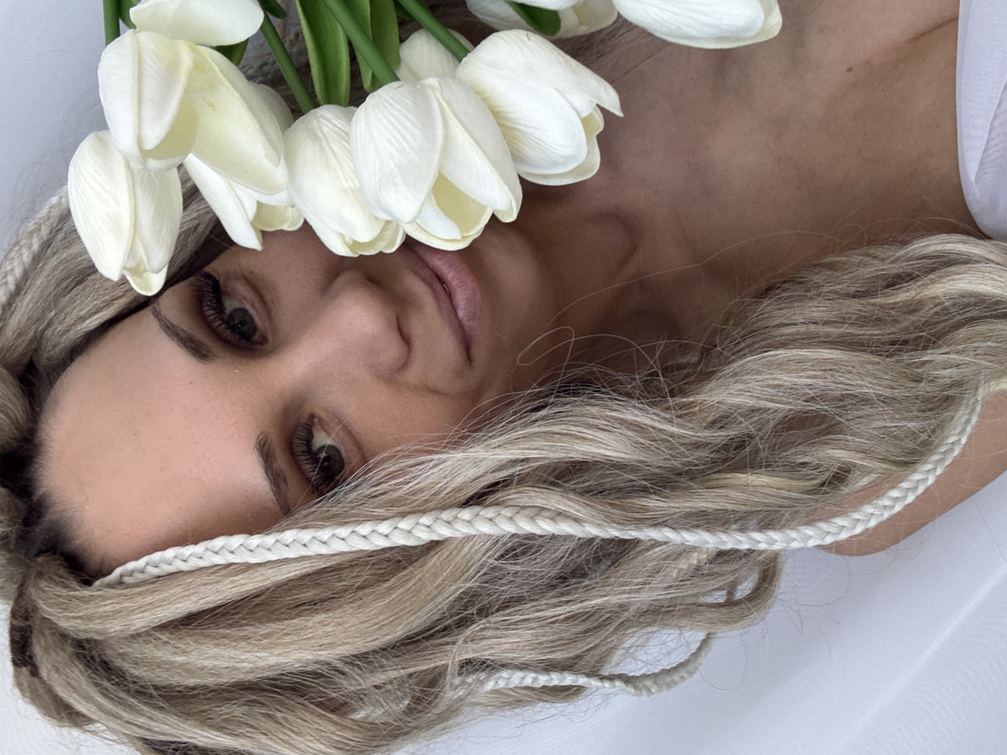 A light-skinned woman with blond hair and braided accents is lying down, head turned toward the viewer, with a bouquet of white tulips partially covering her face. Her eyes are open, and her expression is neutral. She appears to be lying on a white cloth or sheet. It is not possible to confirm the location as Rusanovo, Russia, without additional information.