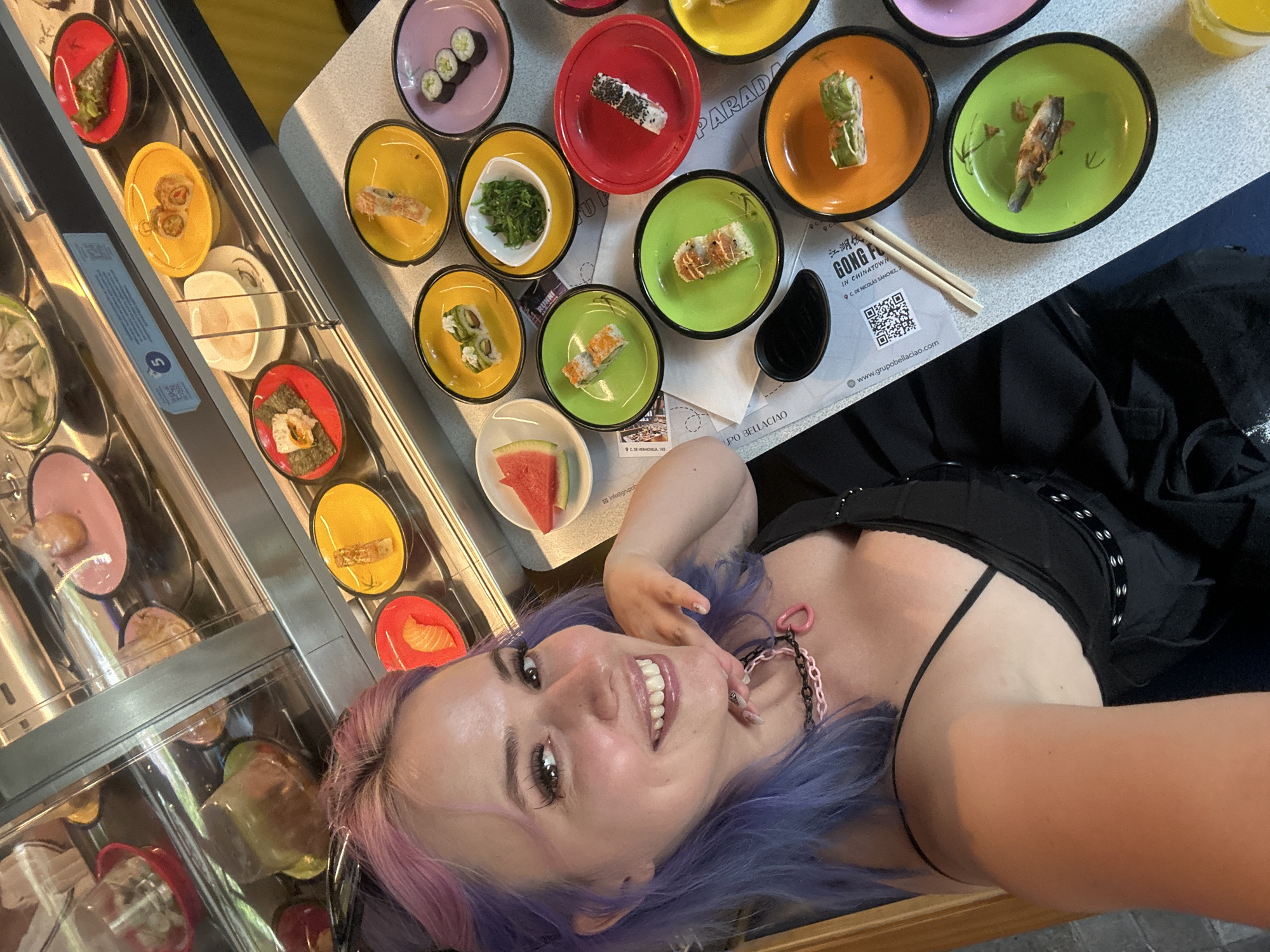 A young woman with lavender-colored hair and a black top is posing for a selfie in front of a conveyor belt sushi restaurant. She is smiling and looking directly at the camera. In the background, various colorful plates of sushi are arranged on a countertop, with a conveyor belt filled with more plates of sushi visible behind her. Chopsticks and a soy sauce dish are also on the countertop. A business card or menu for "Gong Fu Paradiso" in Chinatown on Calle de Nicolas Sanchez 3, is visible. The card also provides another address at C.DE HERMOSALA, 103, which is in the city of Madrid, Spain.