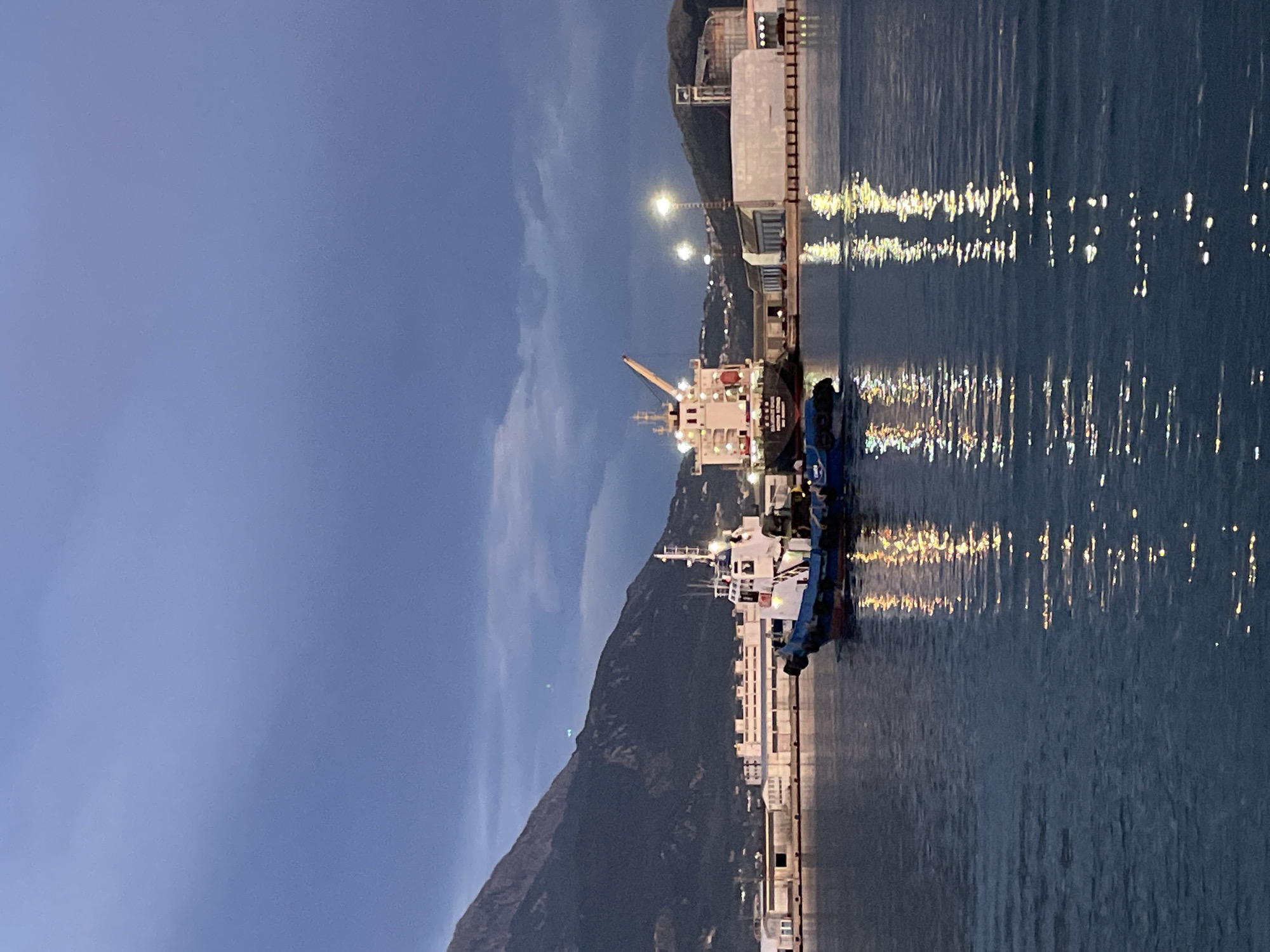 The image shows a harbor scene in Bar, Montenegro, at dusk or early evening. In the foreground are two large ships docked alongside a concrete pier. The pier is illuminated by lights that reflect off the water, creating bright streaks. Behind the ships, a mountain rises steeply, obscuring part of the sky, with a few scattered clouds. Streetlights further up the mountain are also visible. The sky is a gradient of dark blue to a lighter, overcast gray. The overall atmosphere is calm and still, reflecting the quiet of a harbor at night.