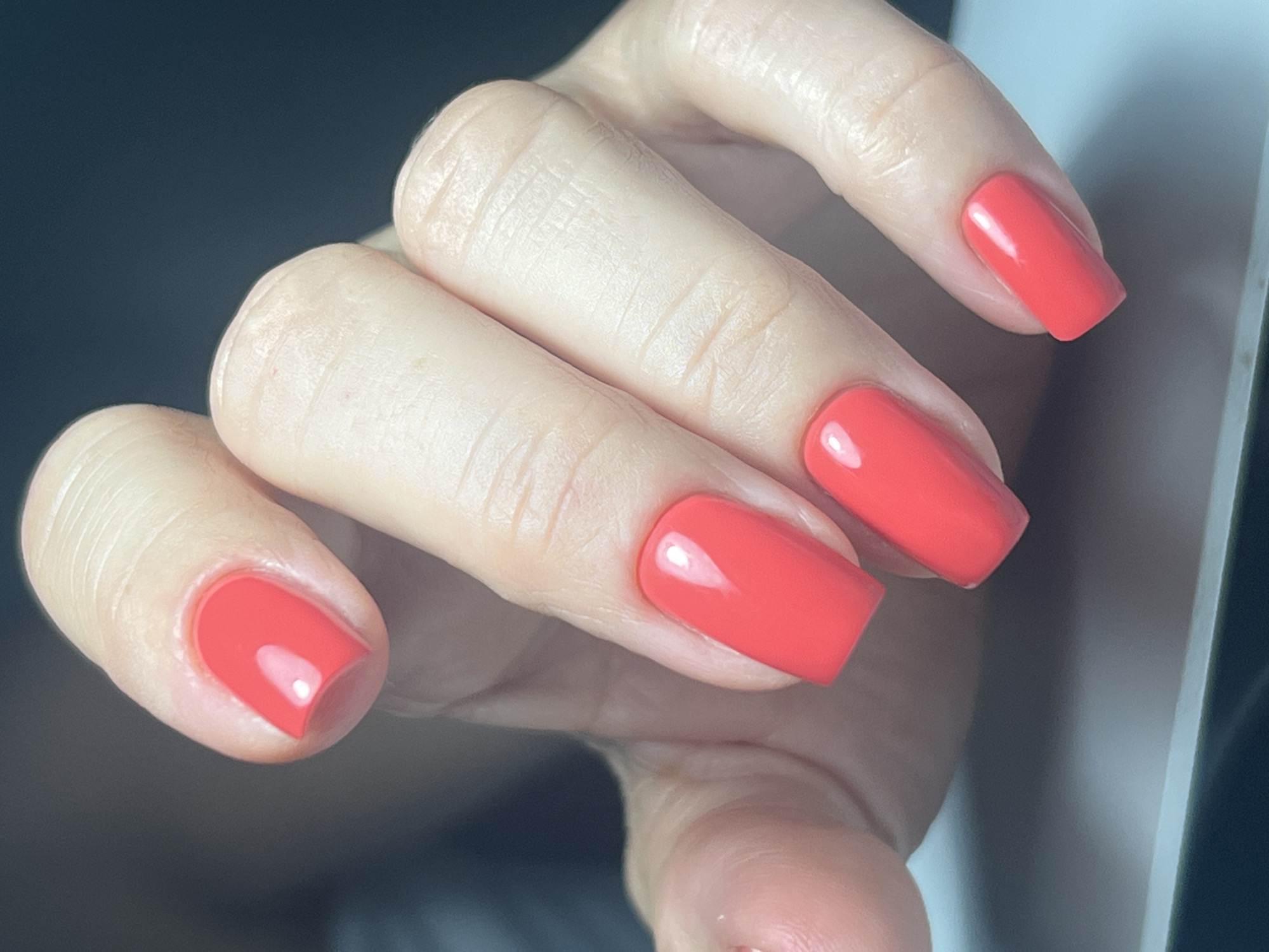 A close-up, high-angle shot focuses on a person's left hand, displaying five square-shaped nails painted a vibrant coral red. The hand is positioned against a dark, blurred background, with a sliver of a light-colored surface, possibly a table, visible at the bottom. The skin on the fingers appears smooth, with subtle creases and natural skin texture. A bright highlight reflects off each nail, indicating a glossy finish. The image is tightly framed, capturing the detail of the manicured nails and the skin surrounding them. No specific location in Athens, Greece, is discernible from the visual information provided.
