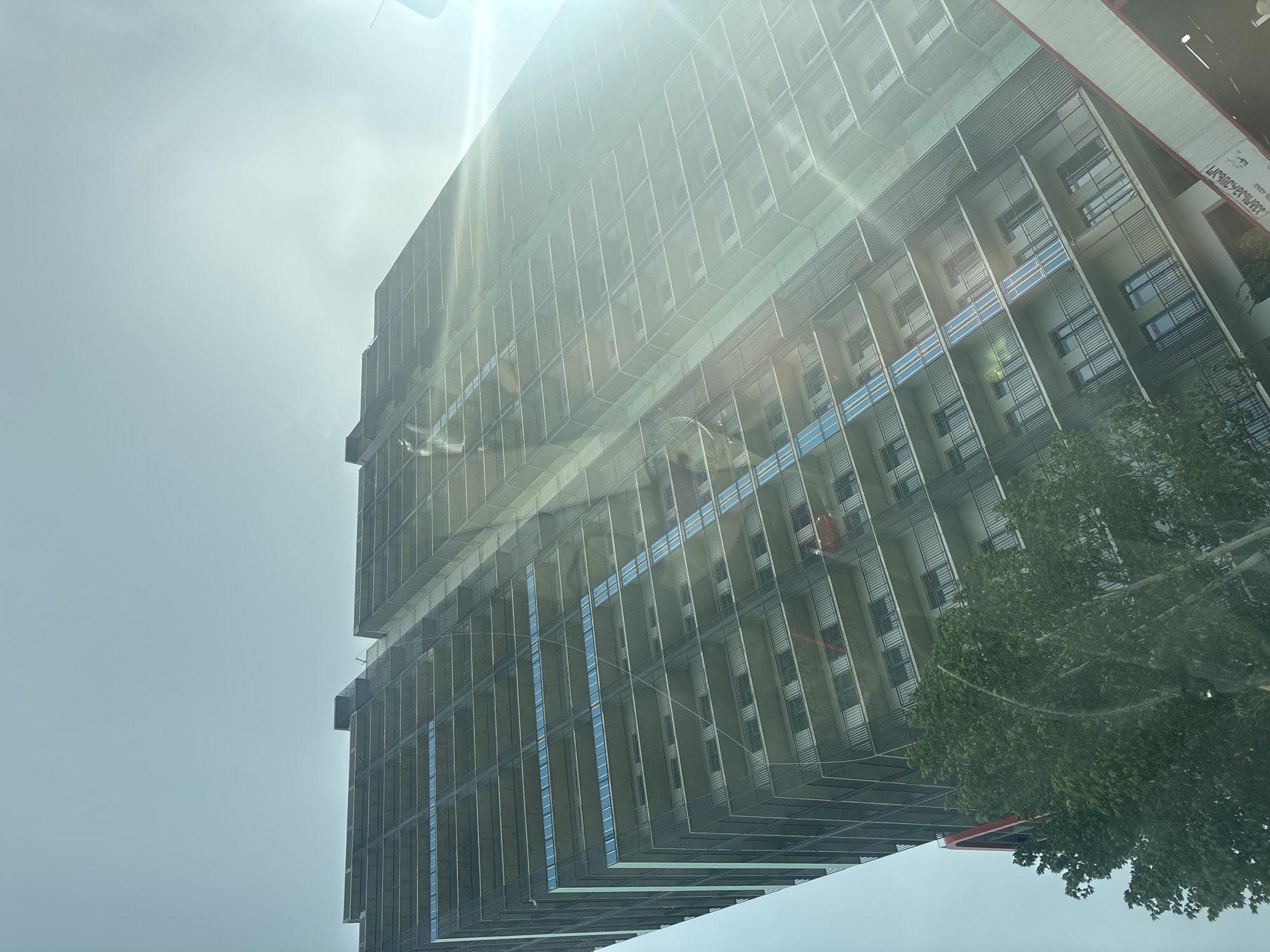 The image shows a tall building under construction, likely in Tbilisi, Georgia. The building is multi-storied with a repetitive pattern of windows and balconies. It appears to be shrouded in scaffolding and netting, suggesting ongoing construction or renovation work. The sky is overcast, and there's a bright glare, possibly from the sun, partially obscuring the top of the building. A tree's foliage is visible in the lower right corner of the image, and there is an inscription on a structure on the right side. The image is taken from a low angle, emphasizing the height of the building.