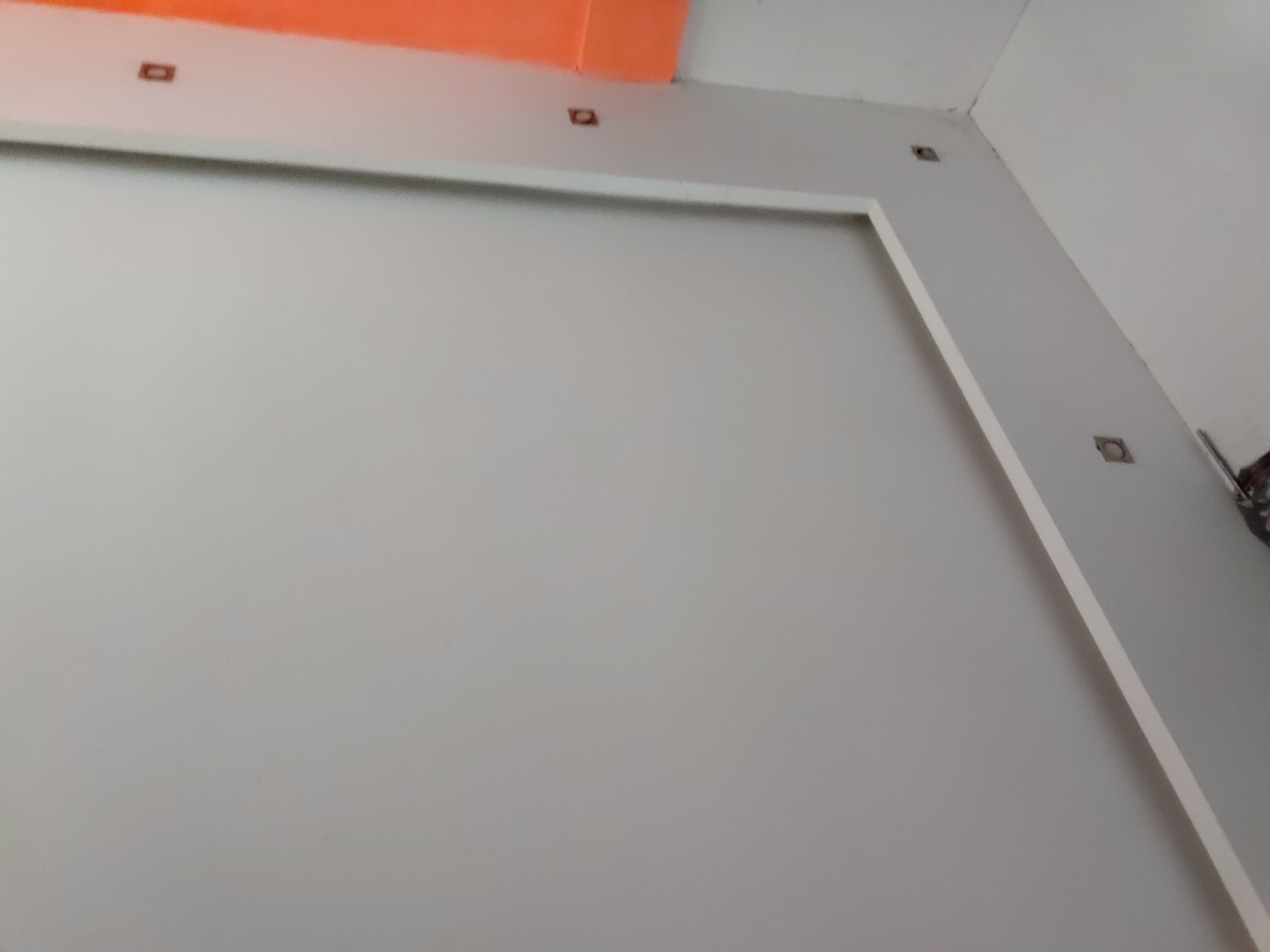 The image presents an interior architectural detail, likely a wall within a room located in Santa Ana, Venezuela. The primary subject is a large, light grey wall, possibly finished with drywall or plaster. A prominent white trim or molding frames a section of this grey wall, creating a defined rectangular or square panel, though only portions of this outline are visible.
Above the grey wall and extending into the upper left corner of the frame, there is a section of ceiling or an upper wall painted in a distinct orange hue, providing a color contrast to the predominantly neutral tones. Along the top edge of the grey wall, within the white trim area, and in the corner where the walls and ceiling meet, several small, square-shaped cutouts are visible. These appear to be pre-drilled holes intended for the installation of recessed lighting fixtures or other electrical components, suggesting that the space is either under construction, undergoing renovation, or awaiting final electrical fittings.
The lighting is even, implying an indoor setting with possibly artificial or indirect natural light. No specific time of day or weather conditions are discernible. The scene is devoid of people and there is no visible text. A small, indistinct dark object, possibly a tool or another fixture, is partially visible in the very bottom right corner. The overall impression is that of a clean, minimalist interior space in an unfinished stage of development.