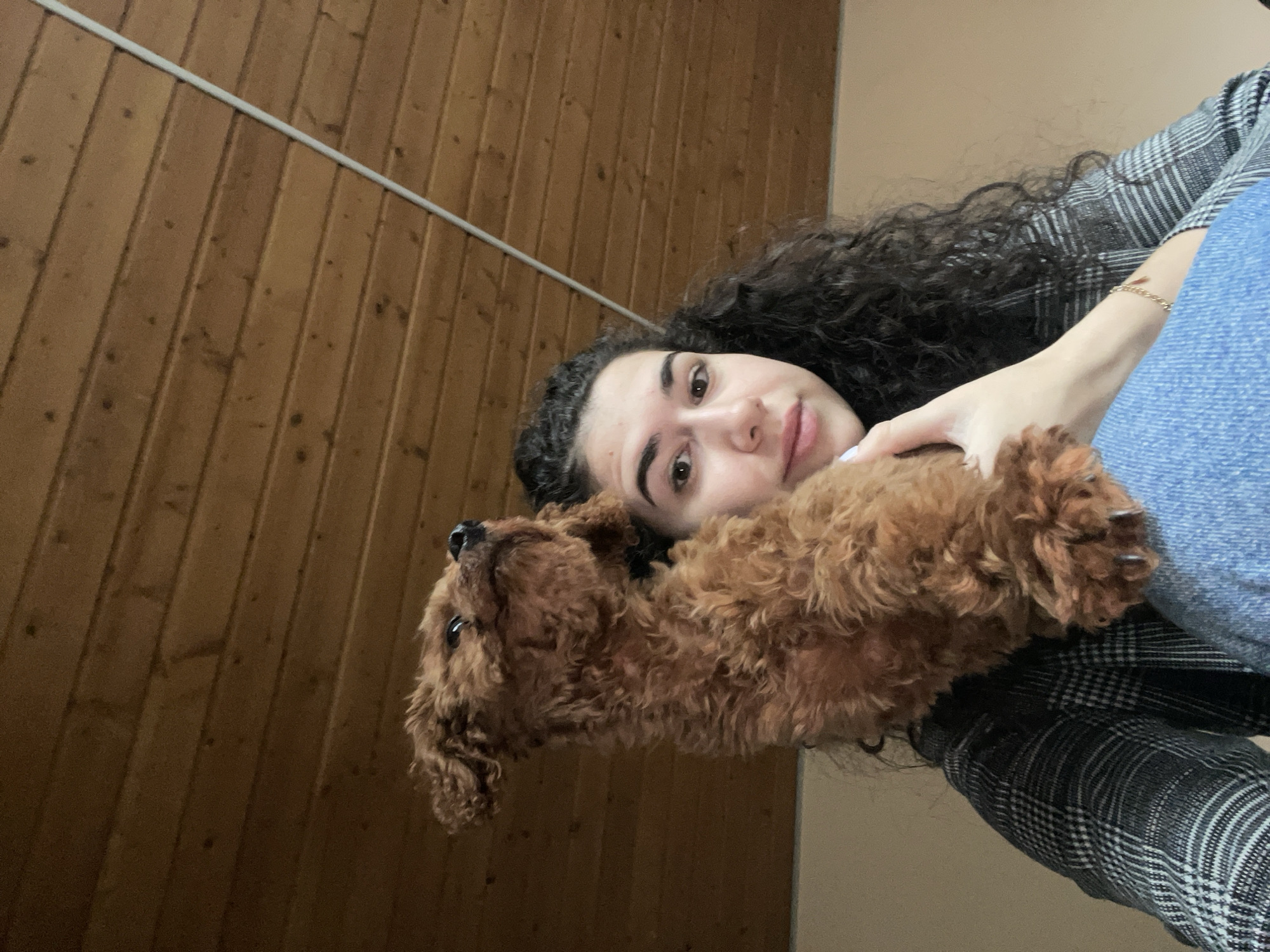 A selfie image shows a woman with dark, curly hair and brown eyes holding a brown poodle. The woman is wearing blue jeans and a black and white plaid jacket. The background is a wooden wall with a white electrical conduit running along the top left. It is difficult to determine if this image was taken in Sofia, Bulgaria.