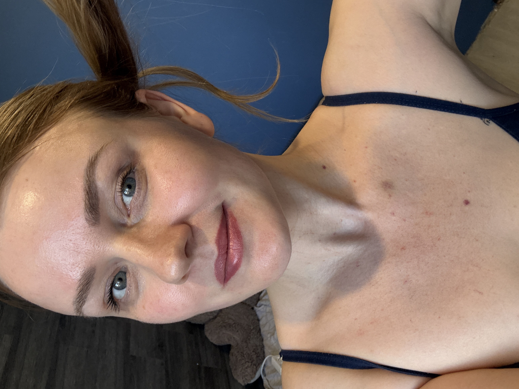 This close-up selfie, taken indoors in Chaikovsky, Russia, features a young woman with light brown hair pulled up, gazing directly at the camera with blue-green eyes and a subtle, gentle smile. Her lips are adorned with pinkish-red lipstick. She is wearing a dark blue or black bra or bikini top, with a strap visible across her shoulder and décolletage. Her skin appears natural, displaying a healthy sheen and some small red marks or freckles on her chest. The background is a plain blue wall behind her head and upper body, transitioning to a dark, possibly wooden surface and a fluffy brown object (like a pillow or stuffed animal) in the lower right portion of the frame. The lighting is soft and even, suggesting an indoor setting during daylight hours. No visible text is present in the image.