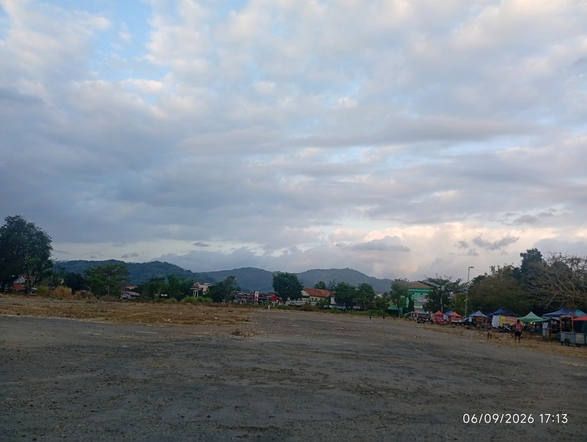 The image captures a wide, open, dusty area in front of a line of buildings and trees, with a mountainous landscape in the background under a cloudy sky. Several tents and stalls are set up along the edge of the open space, suggesting a market or event. There are a few people visible, some walking and some tending to the stalls. The overall scene appears to be a rural or semi-urban area in Purworejo, Indonesia, likely during late afternoon given the timestamp. The ground is dry and dusty, with sparse vegetation. The buildings in the background are varied in style and size. The sky is a mix of blue and grey clouds, with some patches of lighter sky indicating potential breaks in the cloud cover.