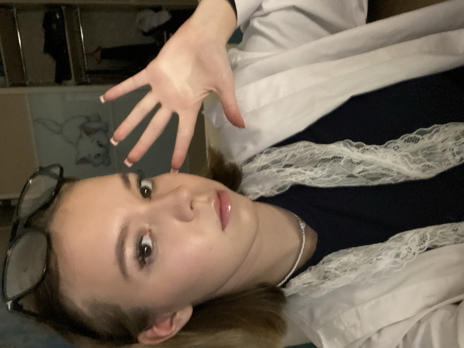 The image presents a close-up, chest-up view of a young individual, likely female, situated indoors in Chaikovsky, Russia. The subject has light brown hair, brown eyes, and is looking directly at the camera with a neutral expression. Black-framed eyeglasses rest on top of their head. A white beaded choker necklace is visible around their neck. They are wearing a dark blue garment underneath an open white collared shirt, which is adorned with a decorative white lace trim. The subject's left hand is raised towards the camera with fingers splayed, displaying light pink manicured fingernails. The background consists of a light-colored wall featuring a graphic of a white cat, and to the right, a dark, partially open wardrobe or closet containing clothing is visible.