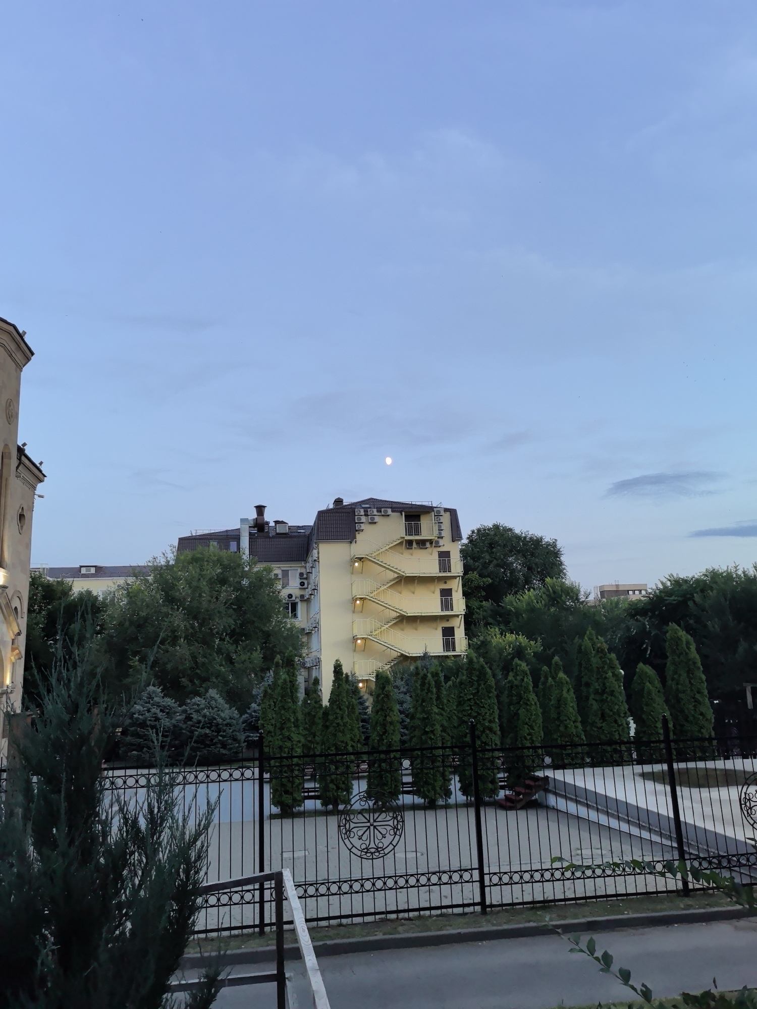 The image shows an outdoor scene in Veshenskaya, Russia, featuring buildings, trees, and a fence. In the upper portion of the image, a pale, full moon is visible against a twilight sky with scattered clouds. The sky transitions from a lighter blue near the horizon to a deeper blue overhead.
Dominating the mid-ground are several buildings. To the left, a portion of a building with beige stone facade and arched windows is visible. Adjacent to it is a multi-story yellow building with dark brown roof. This building has a prominent external yellow staircase winding up its side, with balconies on each level. Several air conditioning units are mounted on the exterior walls of this yellow building. To the right of the yellow building, a taller, darker building with a brown roof and visible air conditioning units is partially obscured by trees.
The foreground is occupied by a black metal fence with an intricate circular design and vertical bars. Behind the fence, a paved area, possibly a courtyard or recreational space, is visible. Several green coniferous trees, some tall and slender and others more rounded, are arranged in rows behind the fence. More mature deciduous trees with dark green foliage are situated further back, behind the fence and in front of the buildings. A dark green, bushy evergreen shrub is in the immediate foreground on the left, partially obscuring the fence. The ground in front of the fence appears to be paved with asphalt.