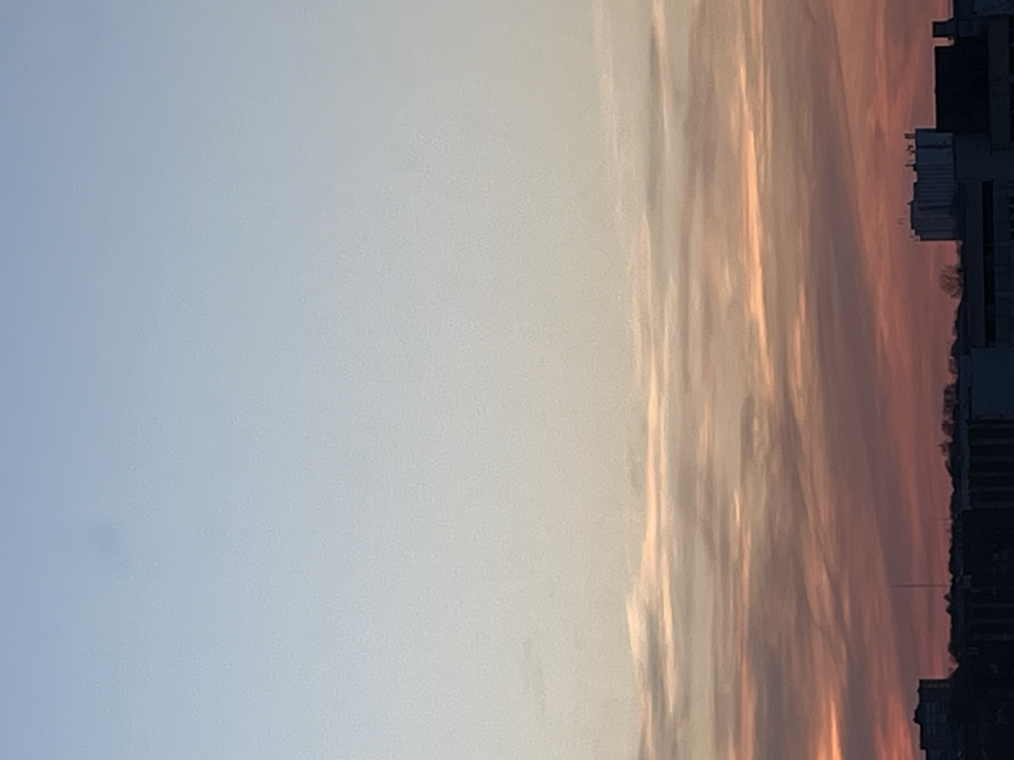 The image displays a cityscape at either dawn or dusk in Izhevsk, Russia, viewed from an elevated perspective. The sky dominates the upper three-quarters of the frame, exhibiting a gradient from light blue at the zenith to pale yellow-orange and then deep orange-red hues towards the horizon. Horizontal stratiform clouds, illuminated by the low sun, are distributed across the middle and lower sky sections. The bottom quarter of the frame reveals a dark silhouette of urban structures. Observable objects include numerous multi-story buildings, appearing as dark masses without specific architectural detail, and sparse clusters of bare deciduous trees. A tall, slender, dark vertical structure, resembling an antenna or communication tower, is visible rising above the cityscape in the central-right area. No individuals or discernible activities are present.