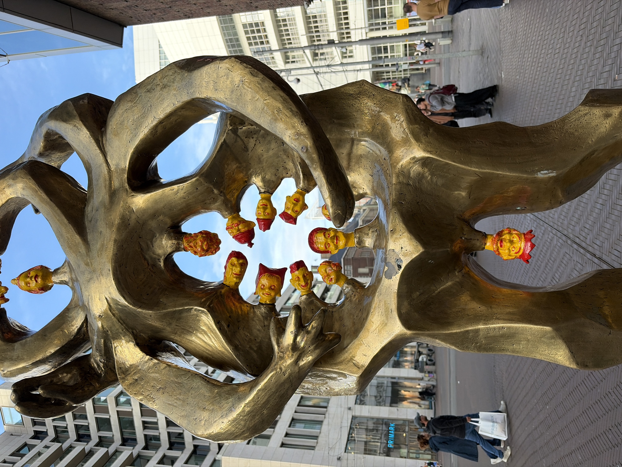 The image captures a large, abstract, golden-bronze colored sculpture positioned outdoors in an urban setting in The Hague, Netherlands. The sculpture features interconnected, irregular forms that suggest stylized human or anthropomorphic shapes, creating several hollow passages. Approximately 15 small, yellow-faced heads with red caps or hair are integrated into these cavities and suspended from various points, including one prominently hanging from the base of the sculpture. Each head displays a distinct, exaggerated, smiling or grimacing expression.
In the background to the left, a tall, modern, light-toned building with numerous rectangular windows is visible. At its street level, a "PRIMARK" retail store entrance is clearly identifiable through large glass windows. To the right, a darker-colored building partially frames the scene, beyond which another multi-story structure with many windows is present. The ground is paved with grey, patterned bricks or cobblestones. The sky above is clear blue with minimal cloud cover, indicating bright daylight.
Several pedestrians are present on the paved area. On the left, a man wearing a grey baseball cap, dark jacket, and blue jeans walks with a woman in a dark blue coat, both moving away from the viewer. The man carries a white shopping bag displaying the "PRIMARK" logo. On the right, multiple indistinct figures are scattered, including a man in a light brown shirt and blue jeans walking away, and other individuals standing or moving in the mid-ground. Streetlights and poles are visible amidst the background buildings.