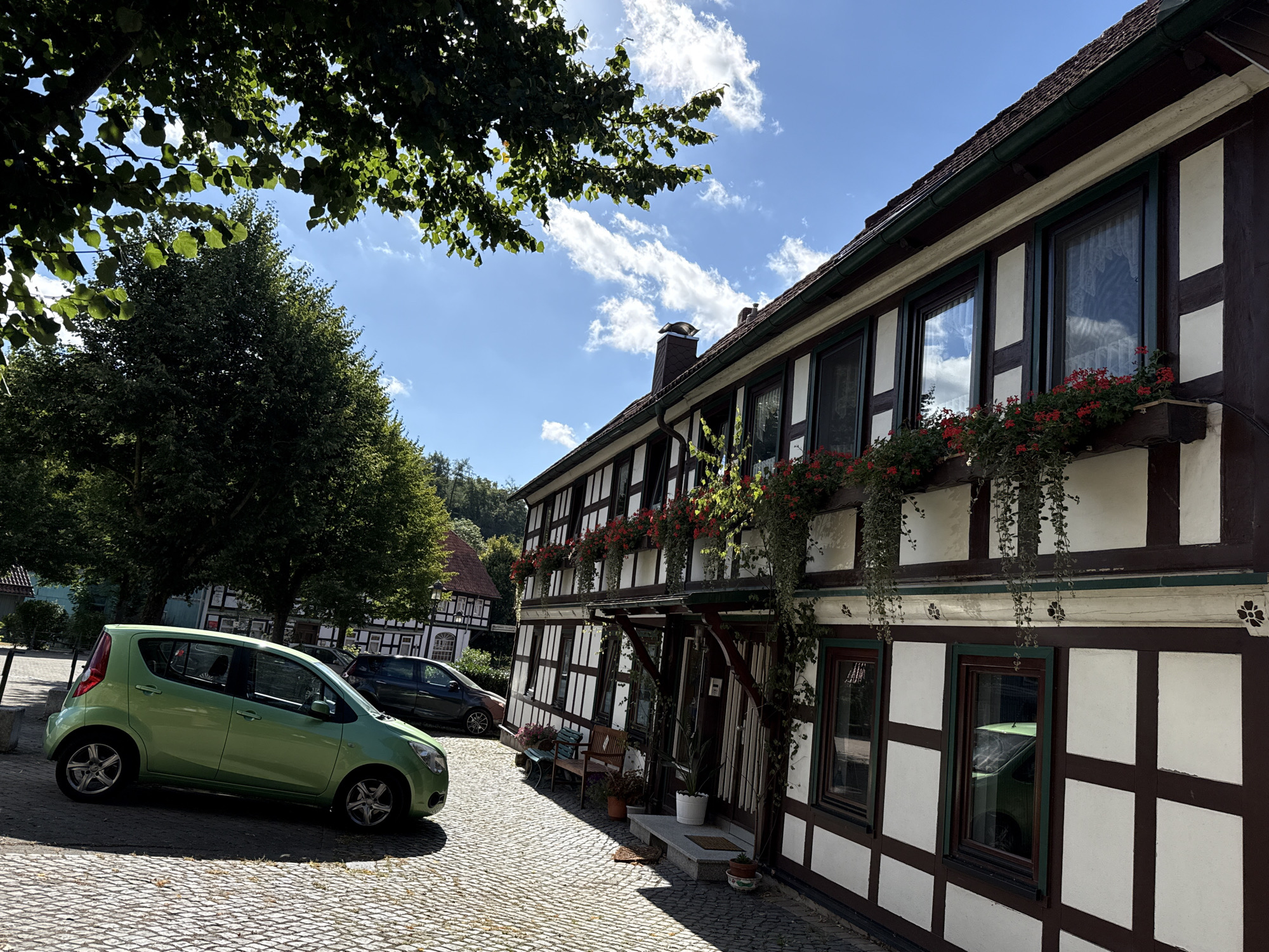 In Walkenried, Germany, a bright green hatchback is parked on a cobblestone street in front of a traditional half-timbered building. The building has dark brown timbers and white infill, with red flowers in window boxes on the upper floors. The scene is bathed in sunlight under a blue sky with scattered white clouds. Trees with lush green foliage are visible in the background. A darker colored car is parked further down the street. The ground is paved with irregular cobblestones.