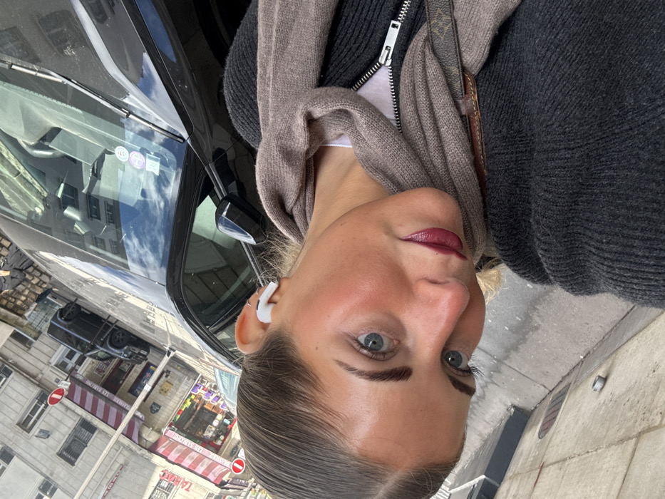 The image, captured at a tilted angle, displays a young adult female posing for a selfie on an urban street. She has slicked-back dark hair, blue eyes, defined eyebrows, and dark red lipstick. A white wireless earbud is visible in her right ear, along with a small hoop earring. She wears a dark grey ribbed zip-up sweater and a light brown knitted scarf, with a white top visible underneath. A brown leather strap featuring a recurring 'LV' monogram pattern is visible over her right shoulder.
The subject is positioned adjacent to a reflective black vehicle, likely an SUV, whose windshield displays several circular French Crit'Air environmental vignettes. In the background, across a street, classic light-colored Haussmannian-style buildings with multiple stories and traditional windows line the street. Below these buildings are storefronts; one features a red awning with "LA TABLE" partially visible, and another displays a red and white striped awning with the clear signage "BRASSERIE LES SABLONS". A dark SUV is parked further down the street, and red circular "Sens Interdit" (No Entry) signs are visible on street poles.
The presence of "BRASSERIE LES SABLONS" explicitly identifies the location as being in or around Neuilly-sur-Seine, a commune immediately adjacent to Paris, France.