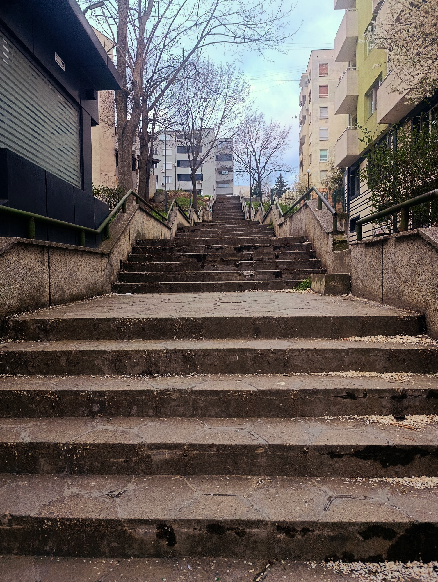 An eye-level shot depicts a long concrete staircase ascending a hill in an urban setting. The steps are worn and partially covered with small, white debris. Metal handrails are visible along the sides of the staircase. Buildings are present on both sides, with bare trees scattered throughout the area. The sky is overcast. The staircase is located in Kardzhali, Bulgaria.