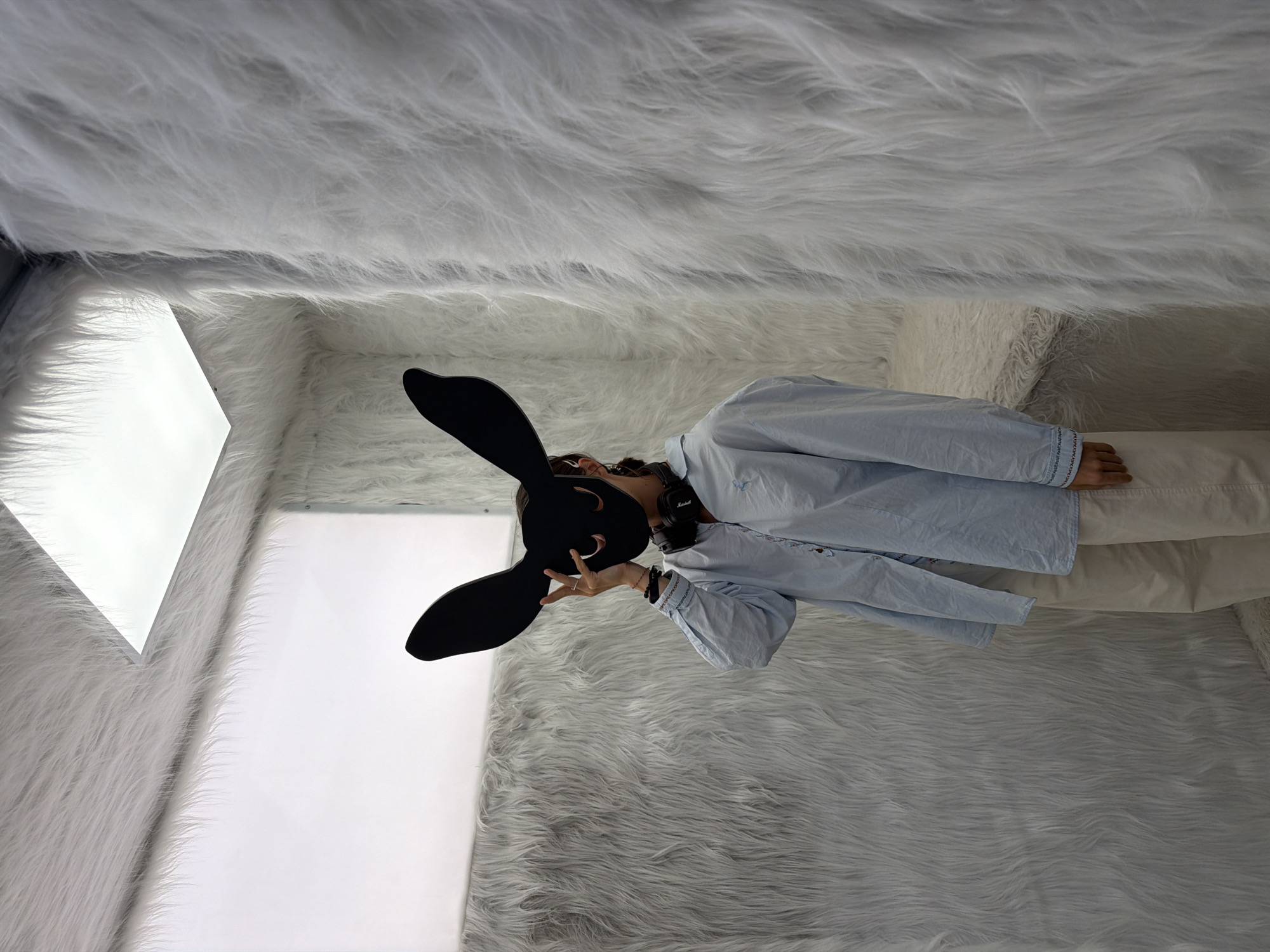 A person wearing a rabbit mask, a light blue shirt, and white pants stands in a room covered with white faux fur. The walls, ceiling, and possibly the floor are completely covered in this material. A bright white light fixture is visible in the corner of the room. The location of this image is unknown.
